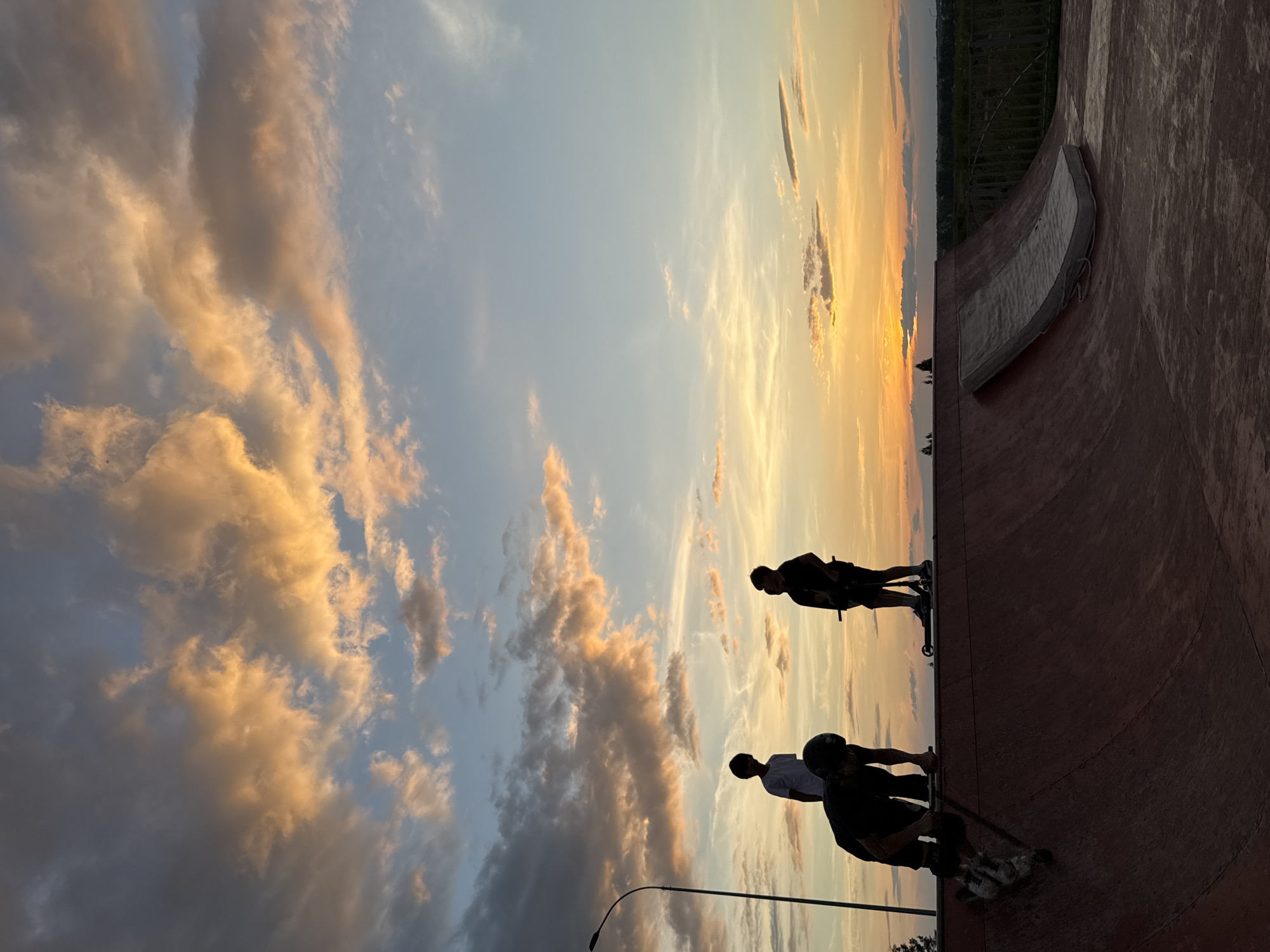The image depicts a skatepark in Mikhaylovsk, Russia, during sunset. Silhouetted figures of individuals are visible against the vibrant sky. In the foreground, a person wearing a helmet is on a ramp, appearing to be in motion with roller skates or inline skates. Further back on the ramp, another individual stands with a scooter, and a third person in a white t-shirt stands looking towards the sunset. The skatepark features a large ramp structure with a mattress placed on a section of it. The sky is filled with dramatic clouds illuminated by the setting sun, casting warm orange and yellow hues across the scene. A lamppost is visible to the left of the frame. The overall environment suggests an outdoor recreational space.