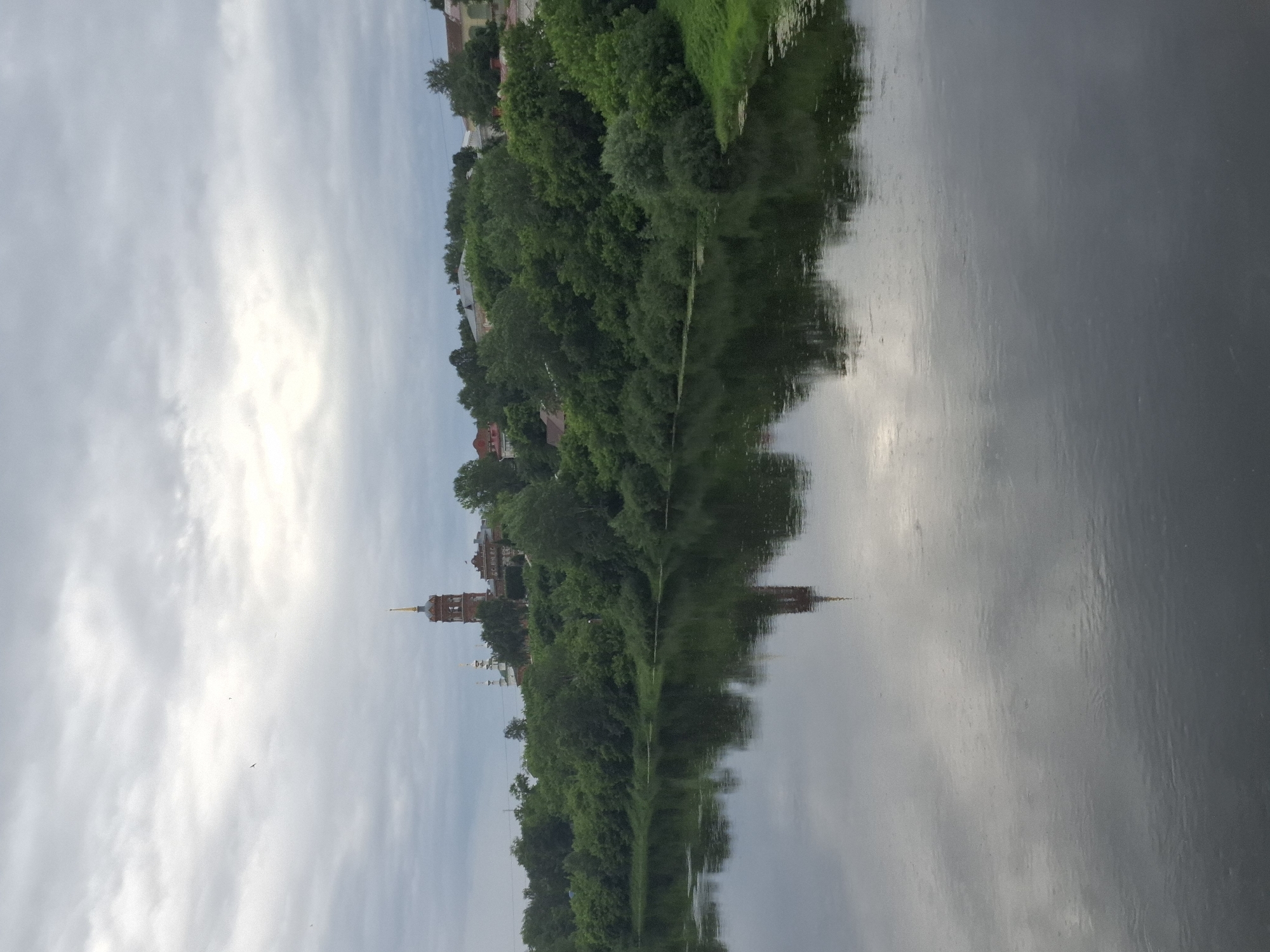 The image captures a scenic view of the Iren River in Kungur, Russia, with a prominent church steeple reflected in the calm waters. The foreground is dominated by the dark, reflective surface of the river, with subtle ripples distorting the mirrored image of the sky and the opposite bank. The middle ground features a lush, green embankment densely covered with trees and vegetation. Beyond the trees, on a slight rise, stands a traditional Russian church with a tall, brick bell tower topped with a golden spire. Several other buildings, likely residential, are scattered among the trees on the hillside. The sky above is overcast with soft, diffused light, casting a muted tone over the entire landscape. The overall atmosphere is serene and tranquil, characteristic of a riverside town on a cloudy day.