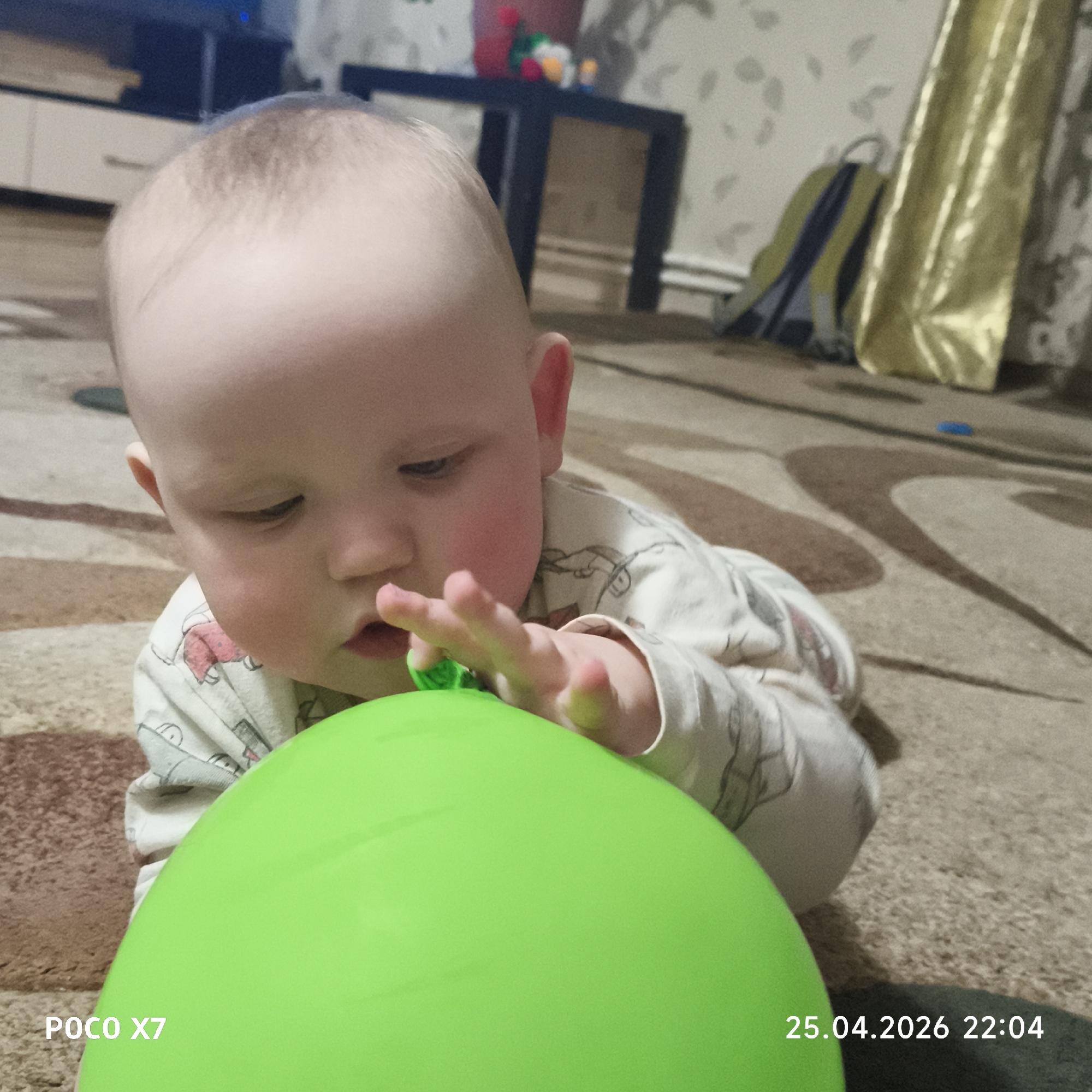 A baby, appearing to be a toddler, lies on its stomach on a patterned brown and beige carpet in an indoor setting, likely a home in Chaikovsky, Russia. The baby has light hair, fair skin, and rosy cheeks, and is wearing a light-colored, long-sleeved onesie or pajama top with subtle patterns, possibly depicting animals.
The primary activity shows the baby keenly engaged with a large, bright lime green balloon positioned in the foreground. The baby's gaze is directed downwards at the balloon, and its right hand is extended, gently touching or grasping the top of the balloon. The expression suggests curiosity and concentration.
In the background, various household items are visible. To the left, a light-colored cabinet or shelving unit is partially seen. Towards the center background, a dark-colored table or stand holds several small, colorful objects. On the right side of the background, a shiny, yellow/gold curtain hangs, and behind it, a blue and yellow backpack rests on the floor. The walls are covered with a light-colored wallpaper featuring a subtle, repetitive pattern.
Notable details include the visible text "POCO X7" in the bottom left corner, indicating the camera model, and the timestamp "25.04.2026 22:04" in the bottom right corner, which indicates the image was captured in the late evening or night. The lighting in the room is artificial.