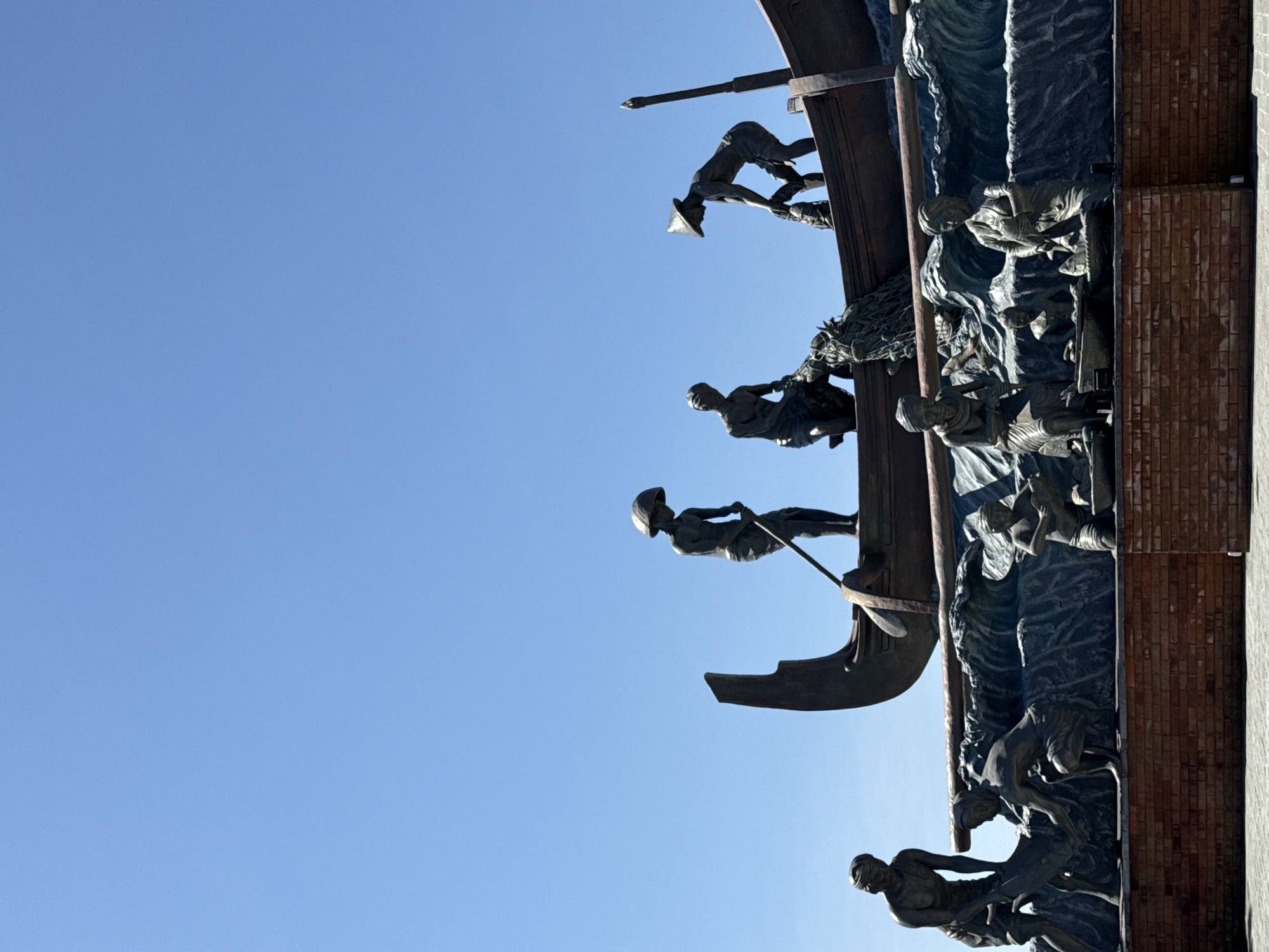 This image presents a detailed outdoor bronze sculpture depicting a traditional fishing and fish market scene, situated in Denpasar, Indonesia. The monument is set against a clear, vibrant blue sky, indicating daylight conditions.
The sculpture is multi-tiered, portraying various stages of fishing. At the highest level, a large traditional fishing boat is depicted, with three male figures actively engaged. One figure, wearing a conical hat, stands with a long oar or pole. Another figure squats, pulling a fishing net filled with fish into the boat, while a third figure, also with a conical hat, bends over to assist with the catch.
Below the boat, two bare-chested male figures on the left are shown hauling a large fish from sculpted waves onto the shore. Further to the right, at a lower level, three figures are involved in processing or selling the fish. A seated female figure with a head covering attends to fish in a basket. Nearby, a smaller figure kneels over another basket of fish. On the far right, a standing female figure, also with a head covering, holds a fish, with additional fish in baskets at her feet.
The entire sculptural ensemble rests on a prominent base constructed of reddish-brown bricks, beneath which a light-colored paved ground is visible. Sculpted waves are intricately detailed around the boat and the figures on the lower levels, enhancing the maritime theme.