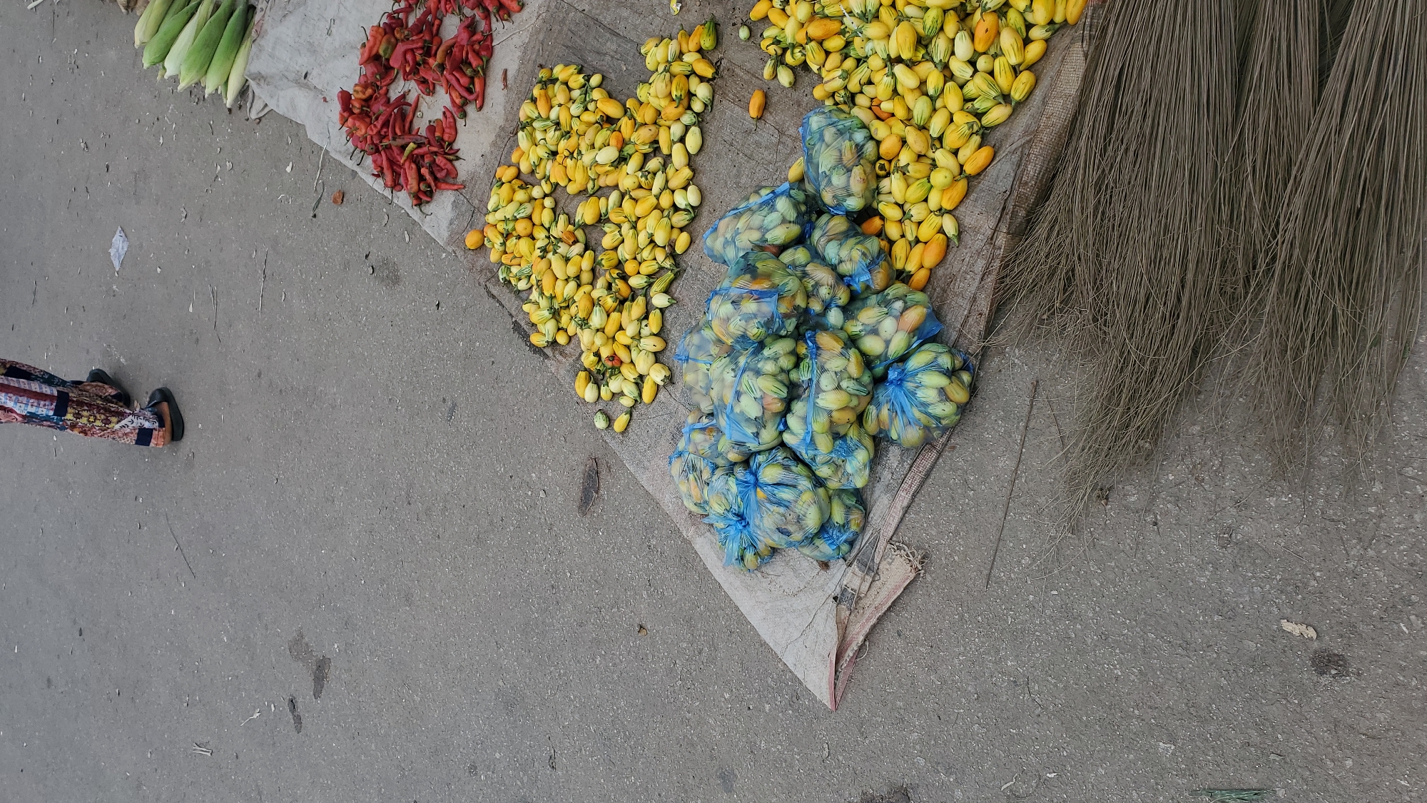 In Jalingo, Nigeria, a street market scene unfolds with produce displayed on a woven mat laid over the asphalt. A person, partially visible from the knees down, stands to the left, wearing patterned trousers and dark footwear, suggesting they might be a vendor or shopper. The market stall features several piles of food items. In the foreground, a large bundle of dried broom material lies on the ground. To the right of the broom, a collection of small, elongated yellow and green fruits, likely a type of pepper or squash, is spread out. Further to the right, another pile of these same small fruits is contained within blue plastic mesh bags, neatly arranged. Beyond these bags, a heap of bright red peppers is visible, and to the far right, several ears of corn are arranged. The ground is paved asphalt, common in urban areas, with scattered debris. The lighting suggests it is daytime.