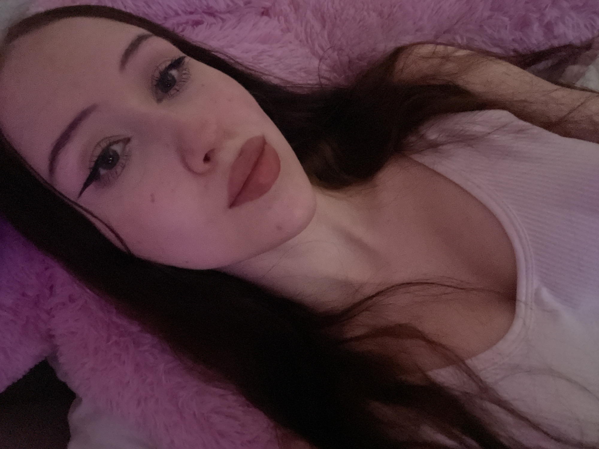 The image is a close-up, high-angle selfie of a young woman with long, dark hair lying on a pink, fluffy surface, possibly a blanket or bed. She is wearing a white ribbed tank top. Her makeup includes prominent winged eyeliner and pink lipstick. The lighting is dim and appears to be colored, casting a pinkish-purple hue over the scene. The background consists of the pink fluffy material and a dark, out-of-focus area. There are no identifiable landmarks or clues that would indicate a specific location in Votkinsk, Russia.