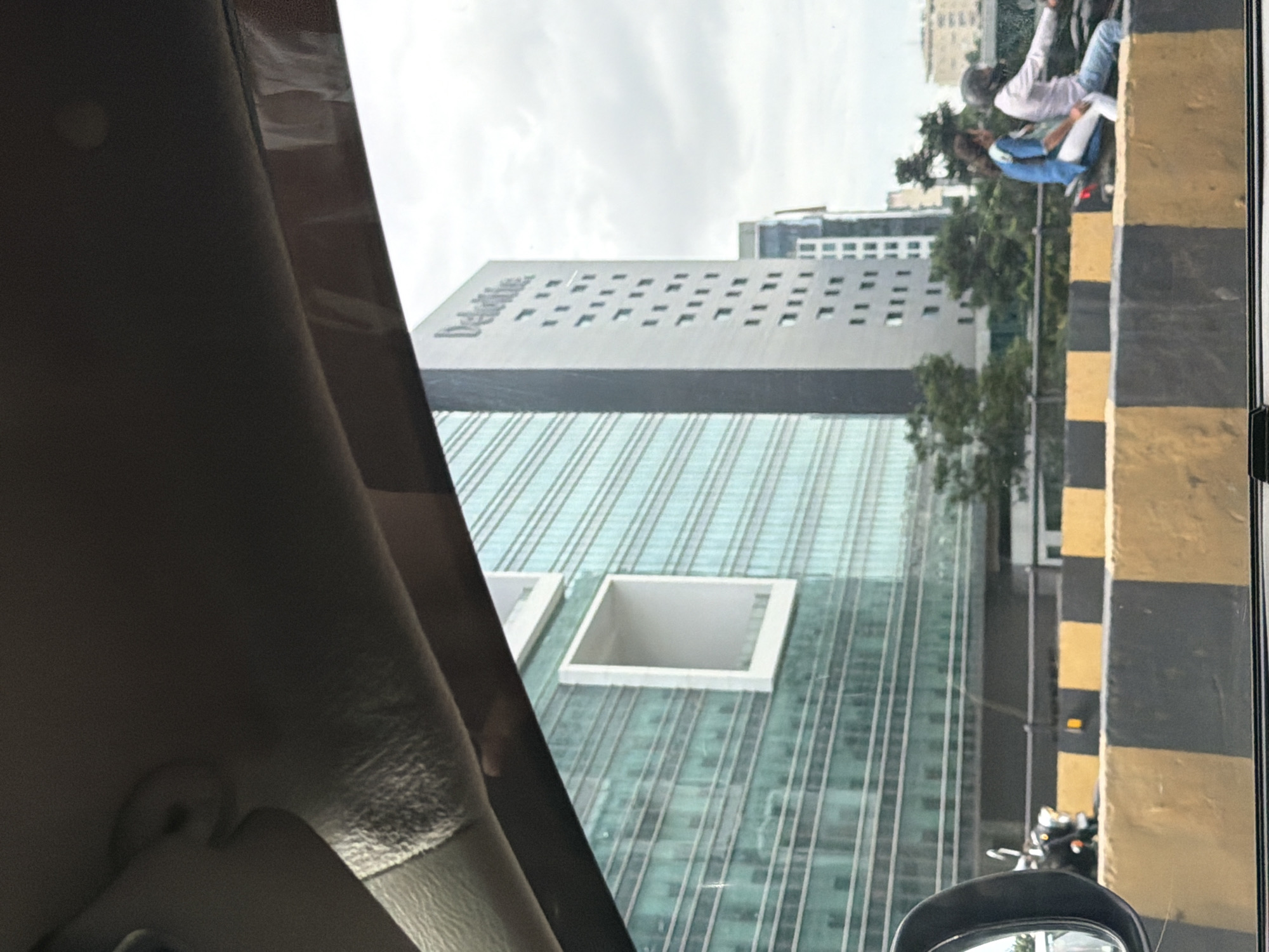 The image captures a street-level view of modern architecture in Hyderabad, India, likely in a business district. In the foreground, a yellow and black striped barrier lines the road, suggesting an elevated highway or flyover. A motorcycle with two occupants, a woman in a blue shirt and white pants and a man in a light-colored shirt and jeans, is moving on the road. The man is wearing a helmet. To the left, a motorcycle is partially visible, appearing to be parked or stopped.
Dominating the mid-ground are two prominent office buildings. The building on the left features a sleek, glass facade with horizontal lines, giving it a modern and reflective appearance. A distinctive, recessed white rectangular cutout breaks the monotony of the glass. To its right stands a taller, darker building with the "Deloitte" logo prominently displayed vertically. This building has a grid of windows. Behind these, other buildings and lush green trees are visible, indicating a well-developed urban environment. The sky is overcast, suggesting a cloudy day. The image is taken from inside a vehicle, as indicated by the car's interior elements visible in the upper portion of the frame, including a portion of the sun visor and the car's interior trim.