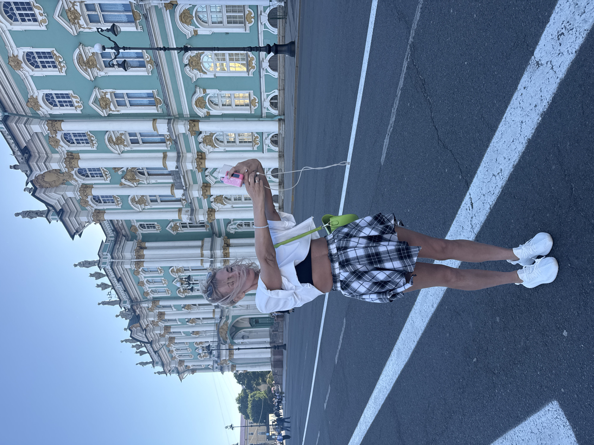 In this full-shot, eye-level photograph taken on a bright day, a woman stands on a street in front of the Winter Palace in Saint Petersburg, Russia. She is facing the camera, holding up a pink smartphone to take a selfie. The woman has blonde hair, pulled back, and is wearing a white cropped t-shirt, a black bra, a black and white plaid mini-skirt, and white sneakers. A bright green purse is slung over her left shoulder. Her right arm is extended, holding the phone and a white cable. The background features the ornate, teal-colored facade of the Winter Palace, which is a significant landmark in Saint Petersburg. The palace is adorned with white columns, gold detailing, and numerous windows. A street with white lines runs across the foreground, and lampposts and trees are visible along the sidewalk. Other pedestrians are visible in the distance on the left side of the frame. The sky is clear and blue.