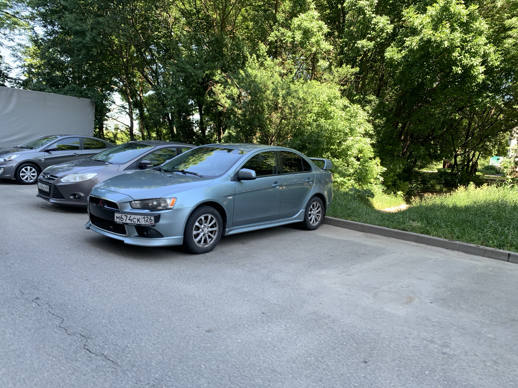 A light blue sedan, identified by its license plate M674CK 126, is parked on an asphalt surface. The car is facing right, with its front bumper and side visible. The vehicle has alloy wheels and a rear spoiler. To the left of the blue car, a dark gray sedan is partially visible, and further left, another gray sedan is parked. The cars are situated in a parking area with a curb and a patch of grass beyond it. Lush green trees with dense foliage form a backdrop, suggesting an outdoor, possibly urban or suburban setting, likely in Stavropol, Russia, given the license plate format. The sky is not visible in the frame. The lighting suggests it is daytime.