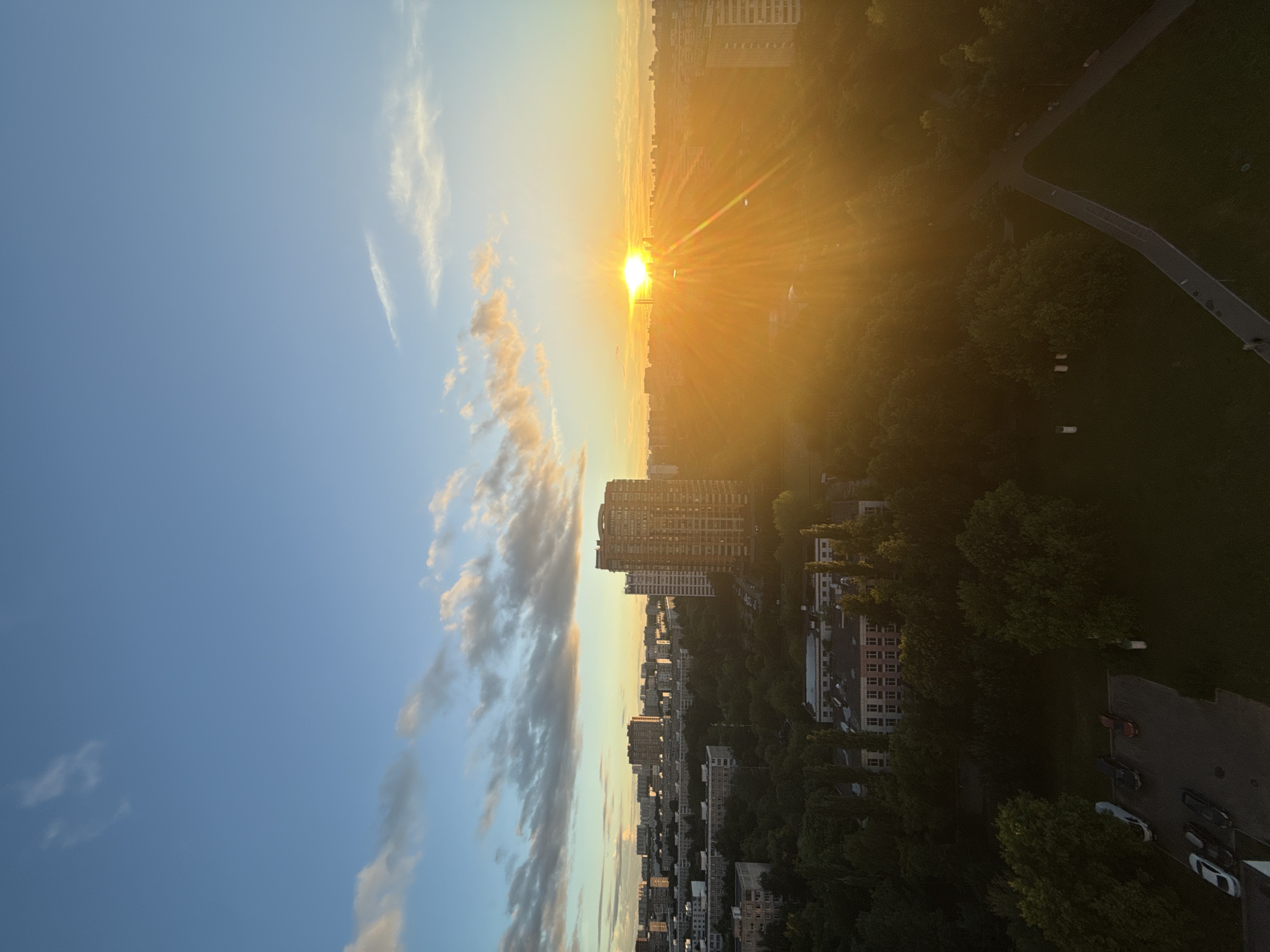 This image captures a sunset over a residential area in Moscow, Russia. The sky is a clear blue with scattered clouds tinged with orange and yellow from the setting sun. A bright sunbeam radiates from the horizon, casting a warm glow over the cityscape. Tall apartment buildings, some modern and some older Soviet-style blocks, are visible amidst lush green trees. A park or green space with walking paths and benches is also present. In the foreground, a parking lot is visible with several cars parked. The overall scene depicts a tranquil urban evening in Moscow.