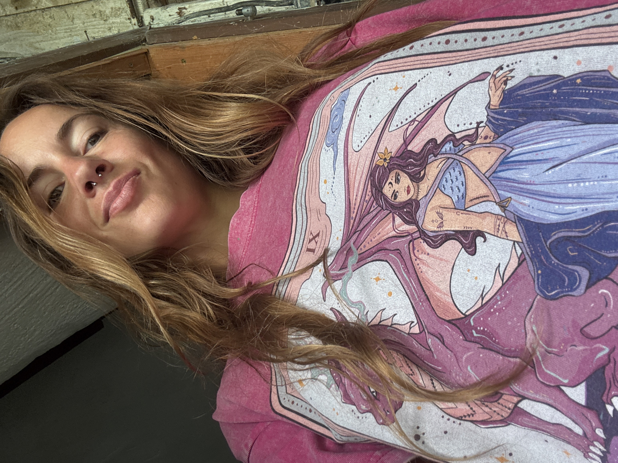 A woman with long, blonde, wavy hair and a septum piercing smiles as she looks down at the camera. She is wearing a pink t-shirt with a graphic print of a dragon and a woman. The print shows a pink dragon with its wings spread, and a woman with long, dark, wavy hair wearing a blue top and a long, blue skirt. The woman in the print has a crescent moon on her forehead. The image appears to be taken indoors, in a dimly lit space, with the background showing a wooden doorframe and a dark wall.