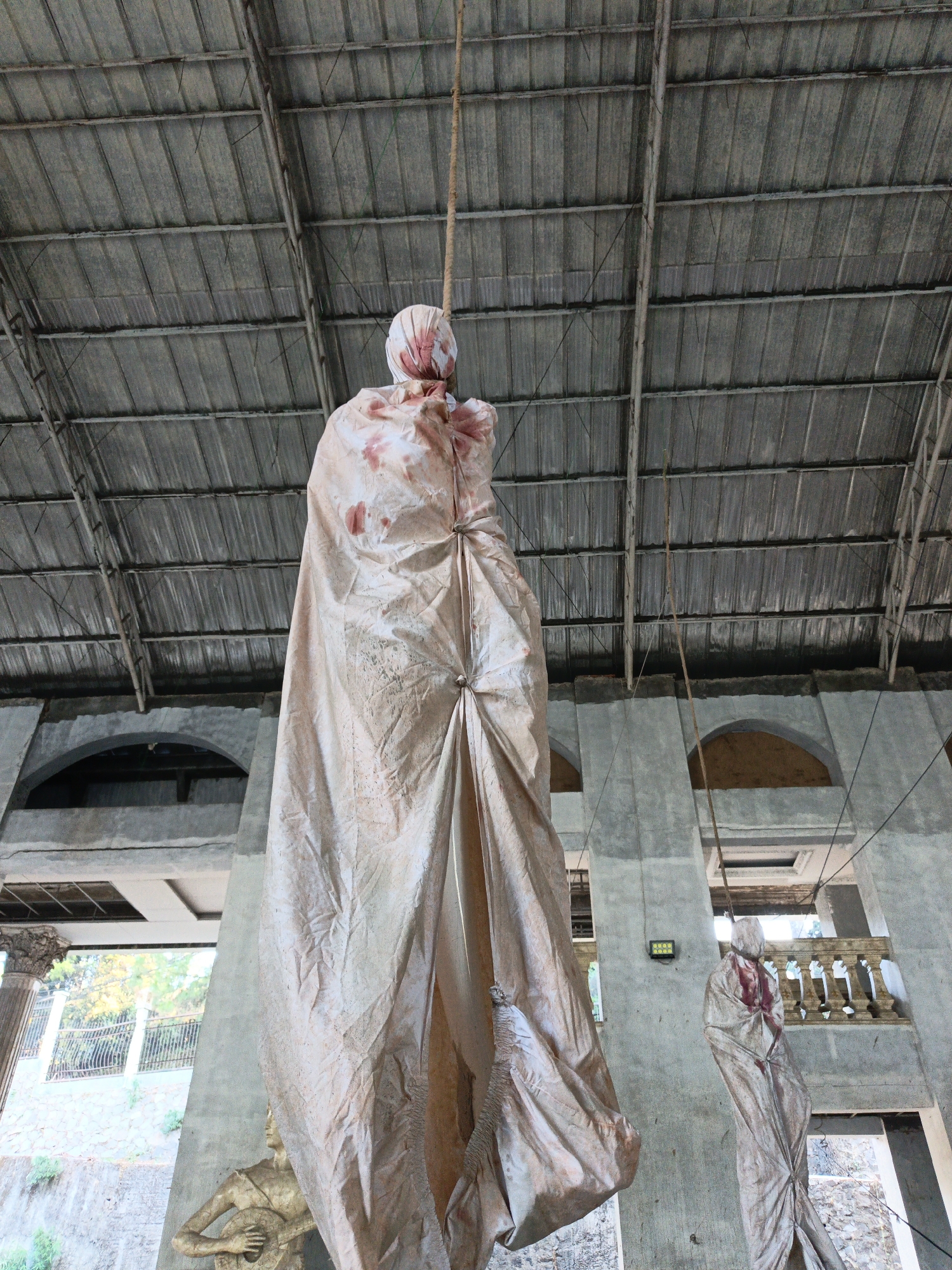 The image depicts an unsettling scene within an abandoned structure. Two effigies, draped in what appears to be a blood-stained cloth, hang from ropes suspended from the corrugated metal roof. The effigies are wrapped from head to toe, with the head area appearing to have some red markings, simulating blood. One effigy is prominently displayed in the foreground, while another is visible further back on the right.

The environment suggests a derelict building with concrete walls and arched openings. Visible structural elements include concrete pillars and a balcony with balustrades. The overall impression is one of decay and disuse.

The location is identified as the abandoned "Rumah Pocong" (Ghost House) in Cibogo, Indonesia. This site is known for its macabre displays, often featuring effigies intended to evoke fear or superstition. The effigies themselves are likely constructed from fabric and stuffing, with the blood-like stains applied for dramatic effect, in line with the typical themes of haunted attractions or horror-themed locations.