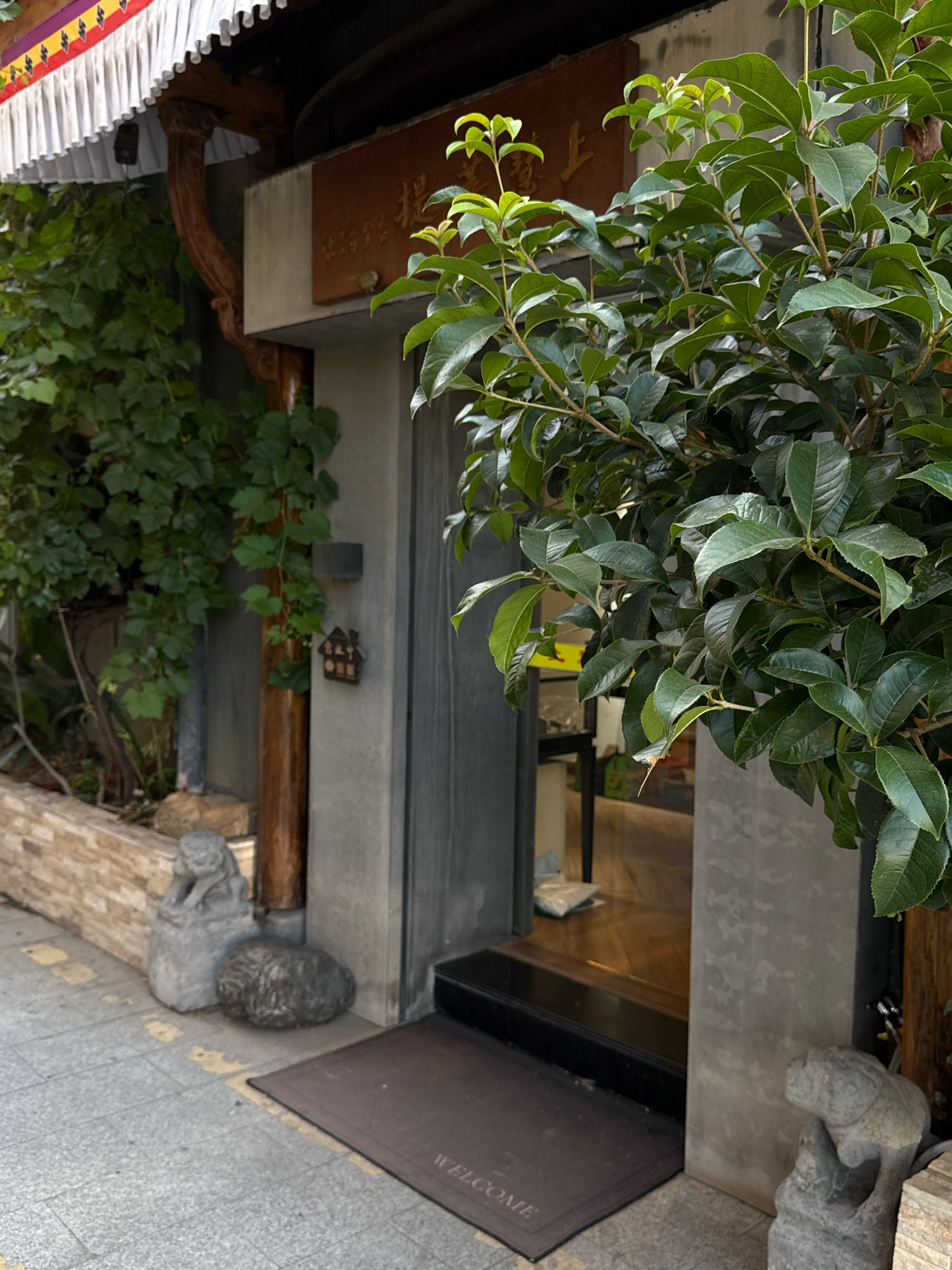 In the city of Unknown, Unknown, an outdoor scene is captured with a building entrance and its immediate surroundings. The facade of the building features a modern design with a grey concrete wall and a dark grey door. Above the door, a brown sign with Chinese characters is visible, partially obscured by lush green foliage. A white valance with colorful tassels hangs above the sign. To the left of the entrance, a section of a brick wall borders a garden area with climbing vines. Two stone statues, one resembling a dog and another a lion, flank the entrance pathway. A dark mat with the word "WELCOME" printed on it lies on the ground in front of the door. The overall impression is of a well-maintained and inviting establishment.