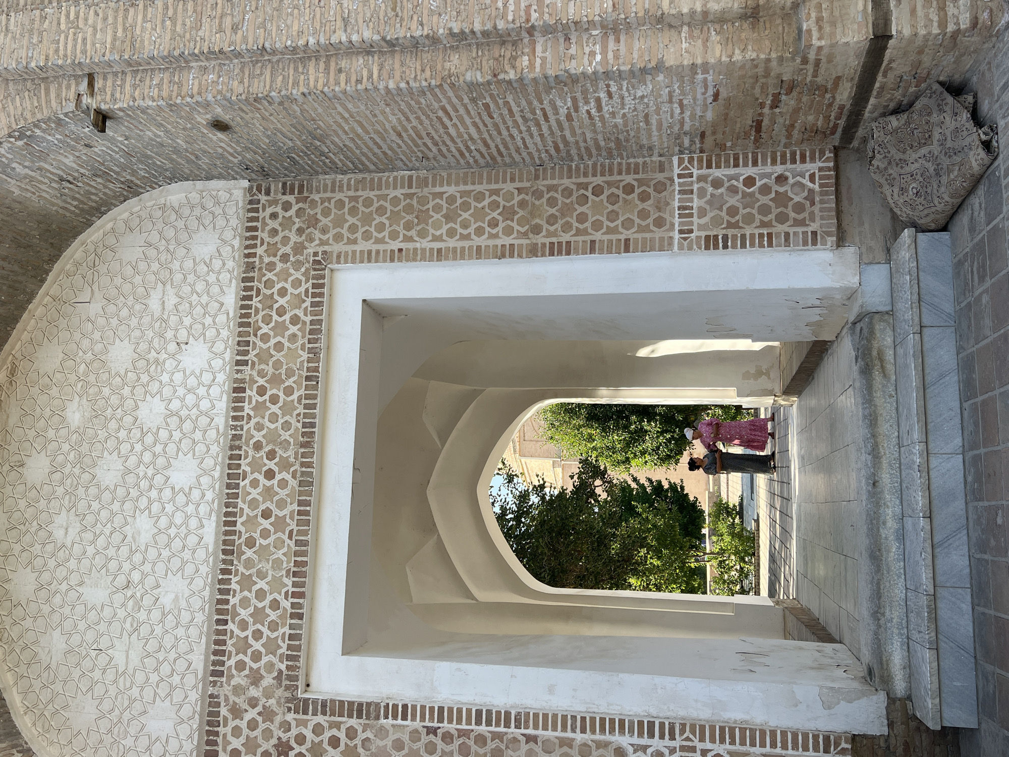 The image shows an ornate archway in Samarkand City, Uzbekistan, leading to a courtyard. The archway is decorated with intricate geometric patterns in light-colored plaster and brickwork. Two women are standing in the courtyard, engaged in conversation. One woman wears a pink patterned dress, and the other wears a dark patterned dress and a white headscarf. They are positioned within a tiled courtyard with trees in the background. In the foreground, a rolled-up carpet is placed against the brick wall next to the archway. The scene is bathed in sunlight, casting shadows and highlighting the architectural details.