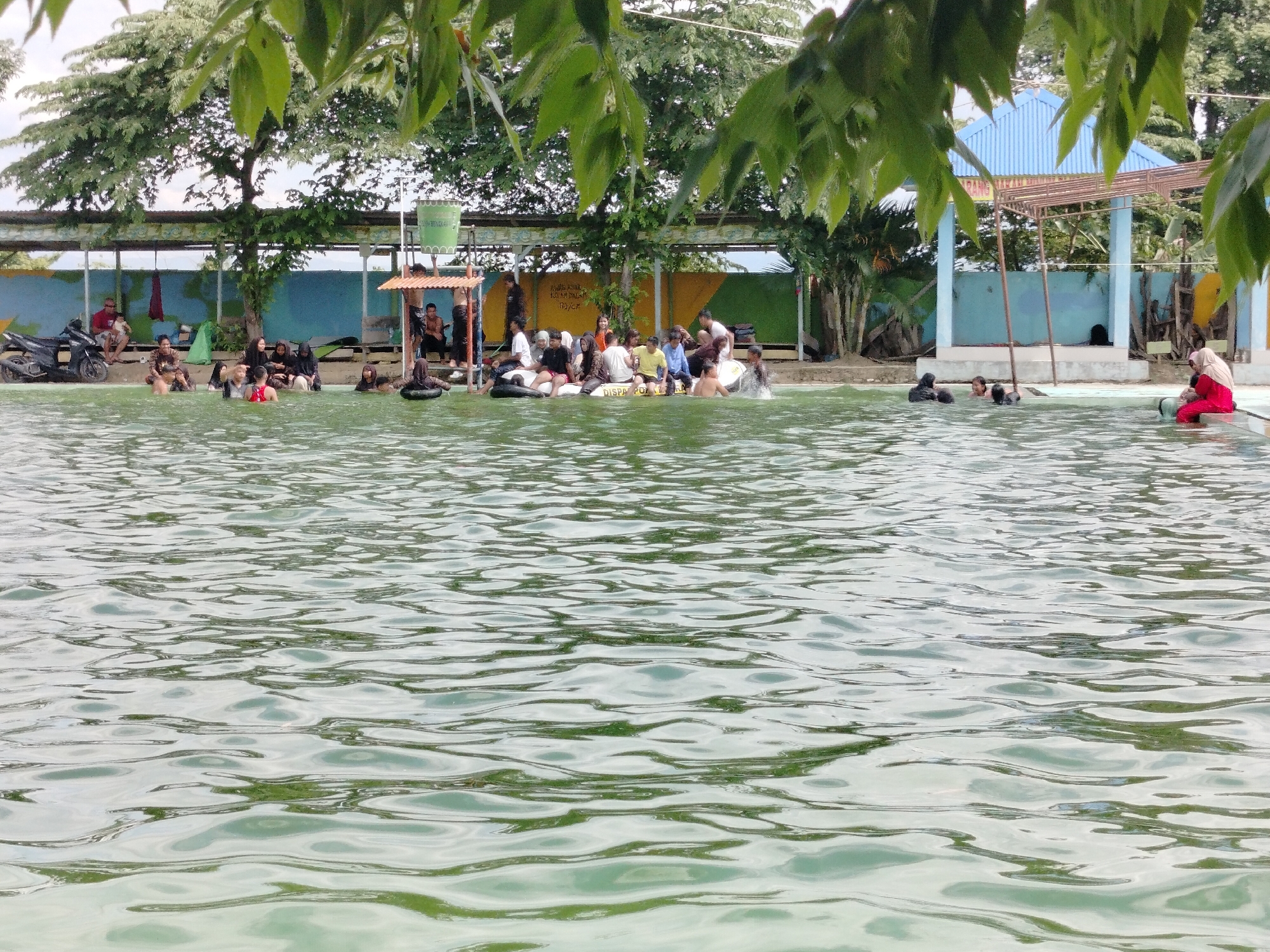 The image depicts a crowded outdoor swimming pool with several people of varying ages wading and sitting in the water. The pool is shallow, with most individuals submerged up to their waists or chests. The background features a blue and white striped awning structure, trees, and a fence. To the left, a black motorcycle is parked near the pool's edge. In the foreground, the water surface is rippled, reflecting the surrounding environment. The scene appears to be a recreational area, possibly a local water park, in Tegarmiko, Indonesia, given the presence of Indonesian text on a sign in the background.