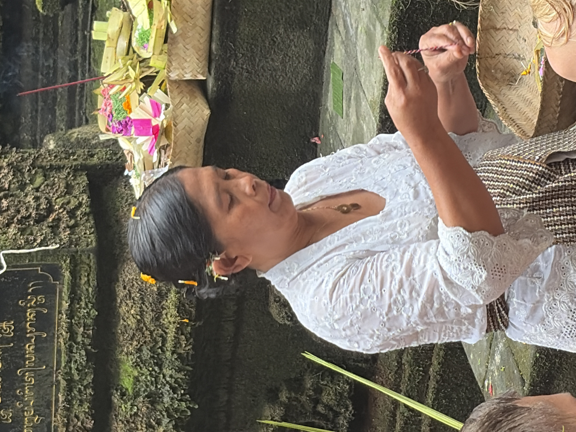 A woman in a white embroidered shirt and patterned sash is seen in Tegallalang, Indonesia, engaged in a ritualistic activity. She holds a thin string, likely for an offering, between her fingers. In the background, woven baskets overflowing with colorful flowers and greenery, along with burning incense sticks, suggest a temple or spiritual site. The woman's hair is adorned with small yellow and white flowers. The environment features moss-covered stone steps and structures, indicative of an ancient or natural setting.