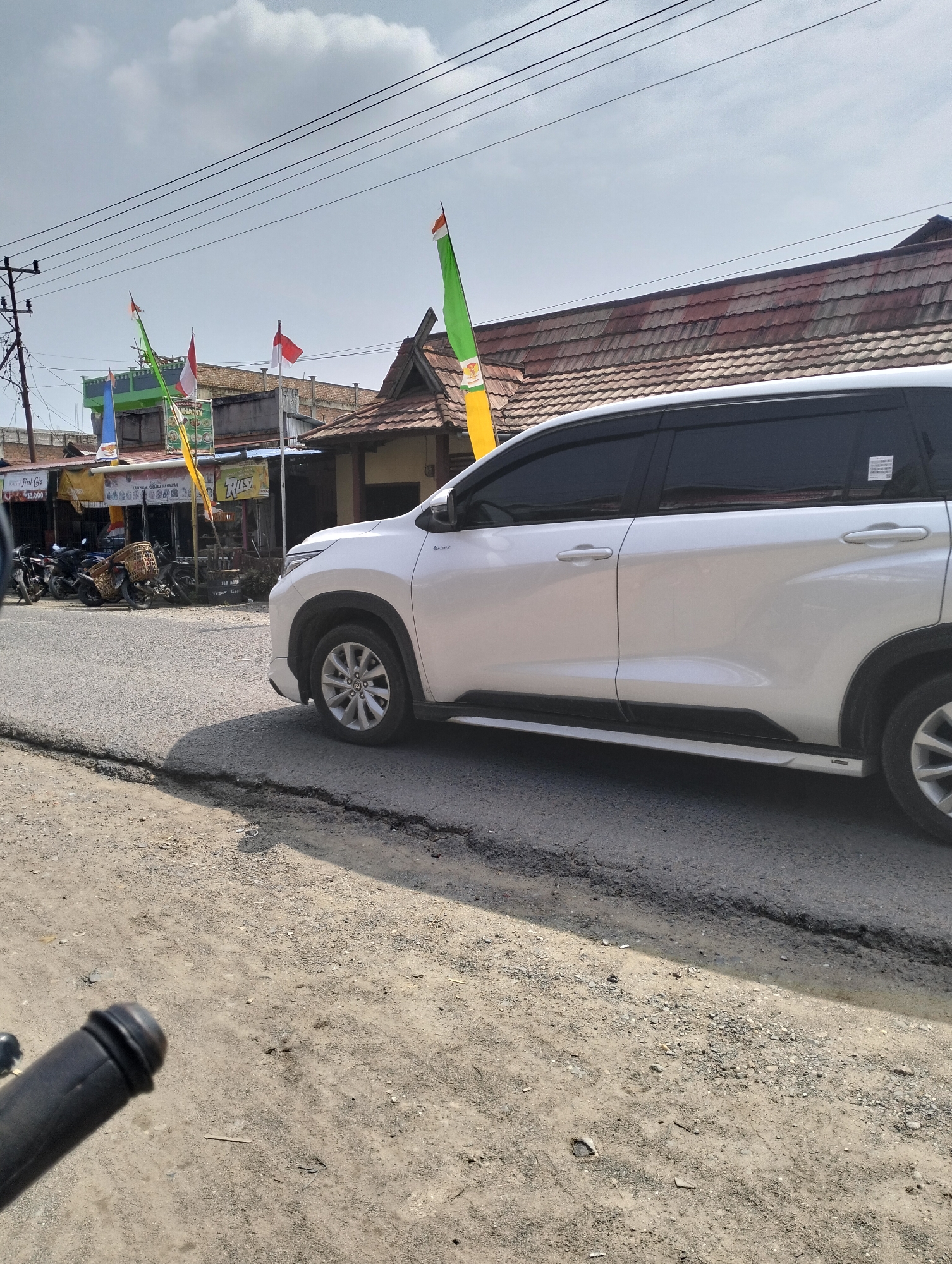 A white SUV is parked on the side of a road in Serdang Jaya, Indonesia. The vehicle is facing right, with its rear end out of frame. In the background, there are several buildings, some with red tiled roofs. Flags are visible, including Indonesian flags, and banners with text in Indonesian. The road appears to be paved but shows some cracking. The area is unpaved on the right side of the road, with dirt and gravel. Power lines are visible overhead. Several motorcycles are parked in front of one of the buildings. The sky is partly cloudy.