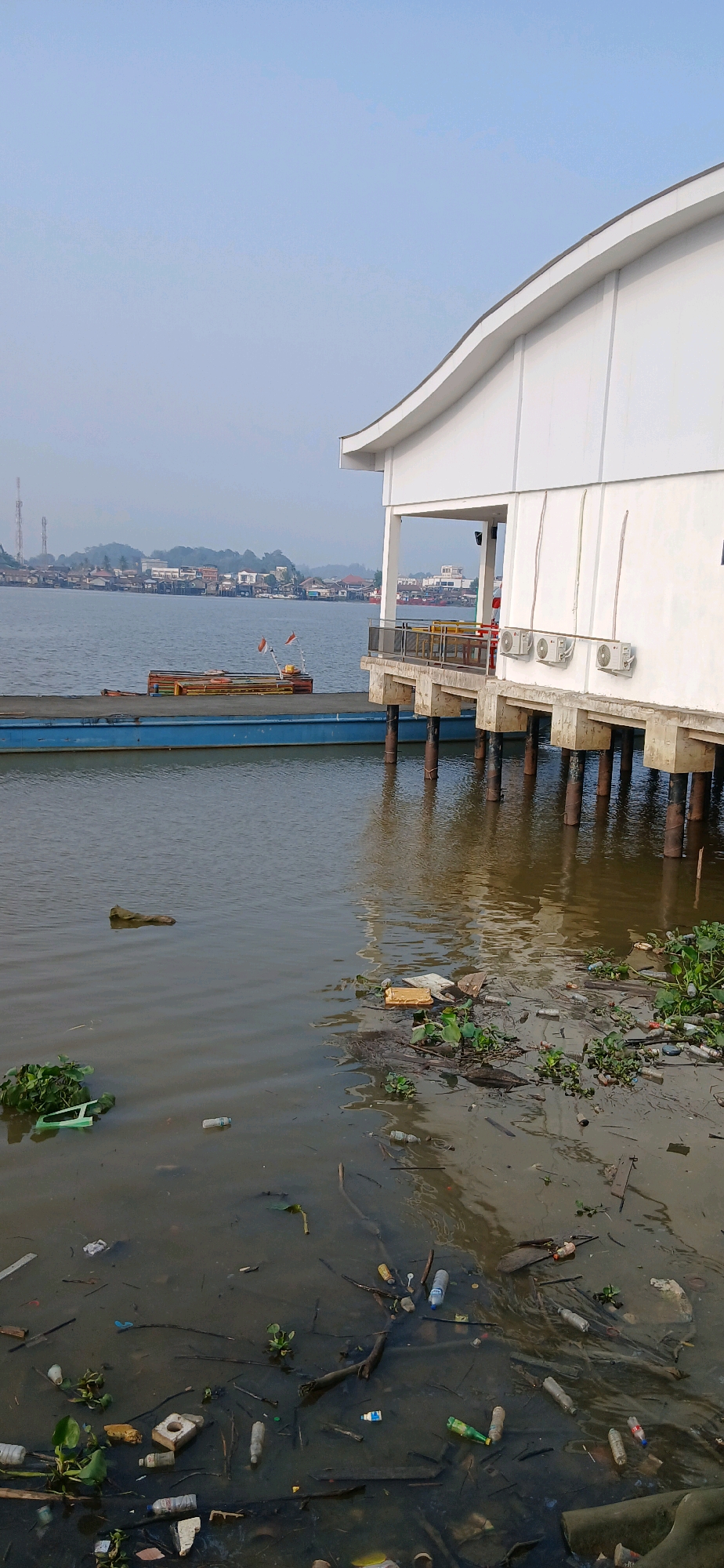 The image depicts a waterfront scene in Samarinda, Indonesia, characterized by pollution. In the foreground, the muddy water is heavily littered with plastic bottles, debris, and aquatic plants. A blue barge is moored in the middle ground, with a smaller boat on top flying two Indonesian flags. To the right, a white building on stilts, likely part of a pier or commercial establishment, extends over the water. The building has several air conditioning units mounted on its exterior wall. In the background, the city of Samarinda lines the opposite bank of the river, with houses and buildings visible, along with some radio towers. The sky is clear and pale blue, suggesting it is daytime.