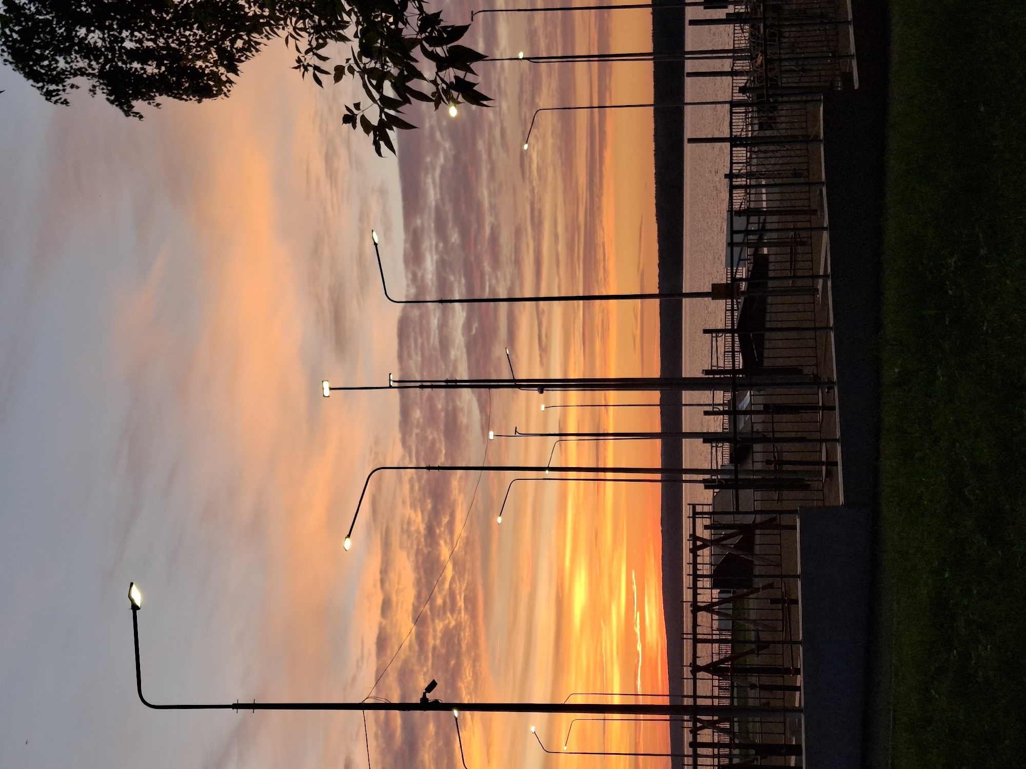 The image captures a serene sunset over the Kama River in Chaikovsky, Russia. A public waterfront area is visible, featuring a children's playground with swings and climbing structures, enclosed by a metal fence. Numerous tall, dark lampposts stand silhouetted against the vibrant sky, their lights beginning to illuminate the dusk. The sky itself is a spectacle of color, transitioning from a bright orange and yellow near the horizon to soft pinks and purples higher up, with wispy clouds scattered throughout. The calm waters of the river stretch out towards a dark, tree-lined embankment on the opposite shore. The foreground consists of a dark grassy area, suggesting a park or recreational space. The overall atmosphere is peaceful and picturesque, marking the end of a day.