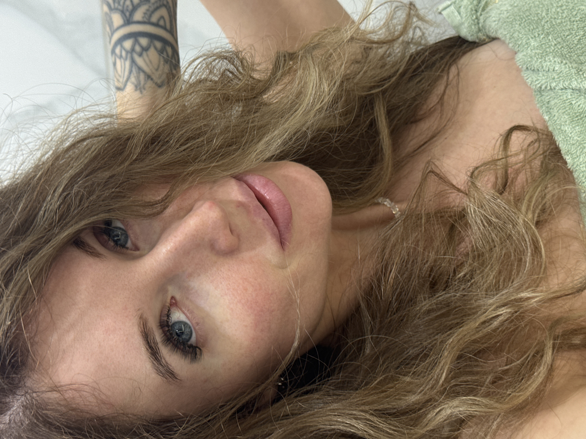 The image shows a close-up, upper body portrait of a woman with long, wavy blonde hair. She has blue eyes, visible eyelashes, and fair skin with some redness on her cheeks. Her lips are slightly parted, and she appears to be wearing light-colored lip gloss. A delicate necklace with small, clear beads is visible around her neck. She has a tattoo on her upper arm, which is partially obscured by her hair and shoulder. She is wearing a light green, textured fabric, possibly a towel or wrap, around her chest and shoulder. The background is a plain, light-colored surface, likely a wall. There is no indication of the city of Chaikovsky, Russia, or any urban environment in the image.
