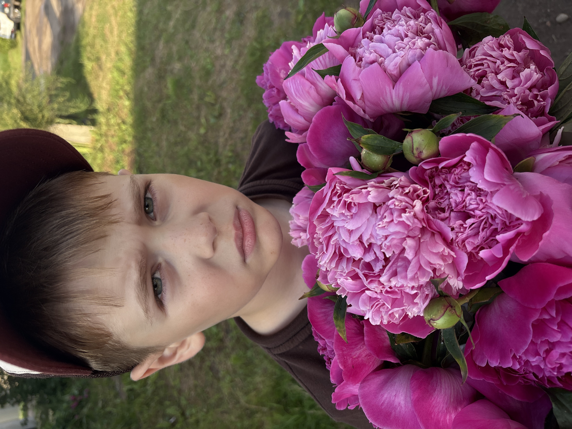 In Niva, Russia, a young boy with short brown hair and freckles is holding a bouquet of vibrant pink peonies. He wears a brown t-shirt and a dark red baseball cap. His expression is a mixed one, with slightly furrowed brows and pursed lips, suggesting a thoughtful or perhaps slightly displeased mood. The background is blurred, showing a grassy area with some trees and a dirt road, indicating an outdoor setting. The peonies are the focal point of the lower half of the image, their rich color contrasting with the boy's attire and skin tone.