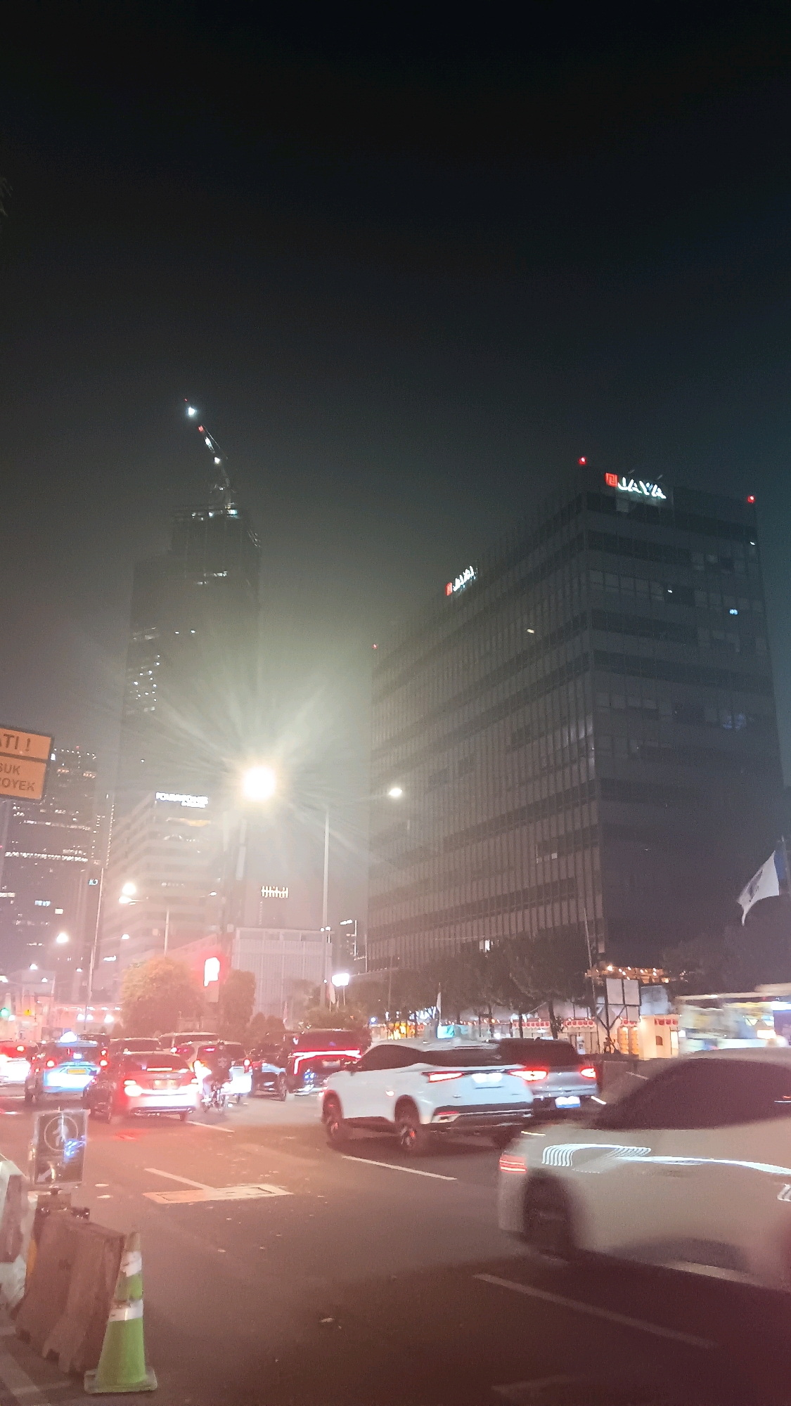 The image depicts a nighttime scene on a foggy street in the city of Central Jakarta, Indonesia, likely in the financial district due to the presence of skyscrapers and office buildings. The foreground shows traffic congestion with several cars, including a white SUV with a blurred reflection of streetlights on its surface, and a white sedan in motion. Cones and barriers are visible on the side of the road. In the midground, more vehicles are present, including a red SUV and a motorcycle. The background features several tall buildings. One prominent building displays a red "JAYA" logo. Another very tall skyscraper, possibly under construction or recently completed, has many lit windows and a construction crane at its peak. Streetlights and building lights illuminate the foggy atmosphere. The overall impression is one of a busy urban environment at night, shrouded in mist.