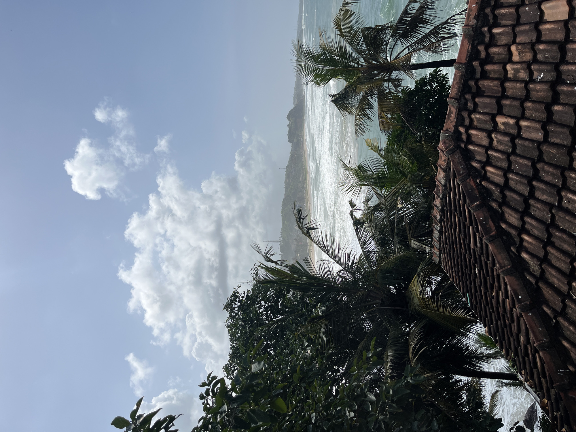 The image depicts a view of the Indian Ocean from a rooftop in Matara, Sri Lanka. In the foreground, a weathered, terracotta tile roof occupies the lower portion of the frame. Beyond the roof, lush green trees and palm fronds frame the scene. The ocean is visible with white-capped waves rolling towards a sandy beach. In the distance, a hilly coastline with scattered buildings and a prominent telecommunications tower can be observed under a sky with scattered white clouds. No people are visible in the scene.