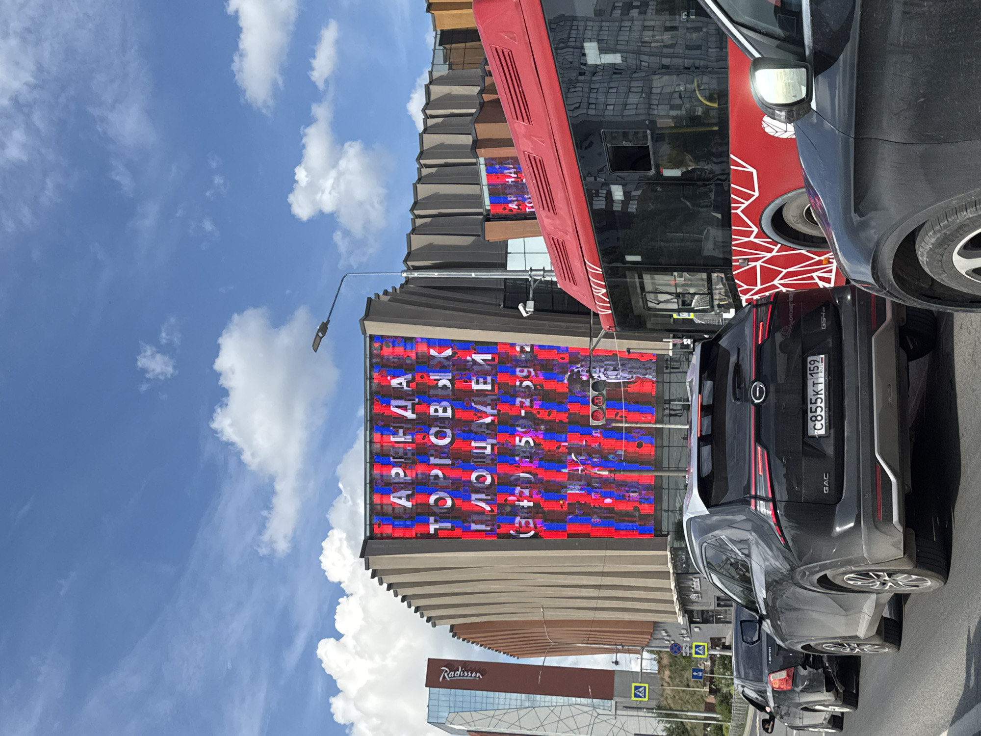 This image depicts a sunny urban street scene in Perm, Russia, characterized by modern architecture and vehicles. The original image is oriented 90 degrees clockwise.
In the center, a striking modern building features a large, vertically oriented LED screen displaying fragmented Cyrillic text. The screen, which appears to be advertising, clearly shows words like "АРЕНДА" (ARENDA - Lease/Rent), "ТОРГОВЫЕ" (TORGOVYE - Trade/Shopping), and "ПЛОЩАДЕЙ" (PLOSHCHADEY - Areas/Spaces), along with numbers like "1-59." The building itself has a distinctive, corrugated or slatted facade in shades of brown and grey, with large glass windows reflecting the sky. To the lower left, another building with a prominent "Radisson" sign is visible, indicating a hotel.
The foreground is dominated by several vehicles. A dark grey GAC SUV is parked or moving slowly, showcasing its rear view with the brand logo "GAC" and a Russian license plate "C855KT 159 RUS," confirming its registration in Perm Krai (region 159). Behind it, parts of another dark vehicle are visible. To the right, a large red vehicle, possibly a bus or truck, is partially seen, with its side adorned with a white geometric pattern. Reflections of buildings are visible on its dark windows.
The sky is bright blue with scattered, fluffy white clouds, suggesting clear weather. The lighting indicates it is daytime, likely midday or early afternoon. A street lamp extends from the left side towards the main building, and part of a traffic light displaying a red signal is visible near the top right of the LED screen. Yellow diamond-shaped road signs are also discernible on the far left, possibly indicating pedestrian crossings or other road information. The overall scene captures a vibrant, modern city environment.