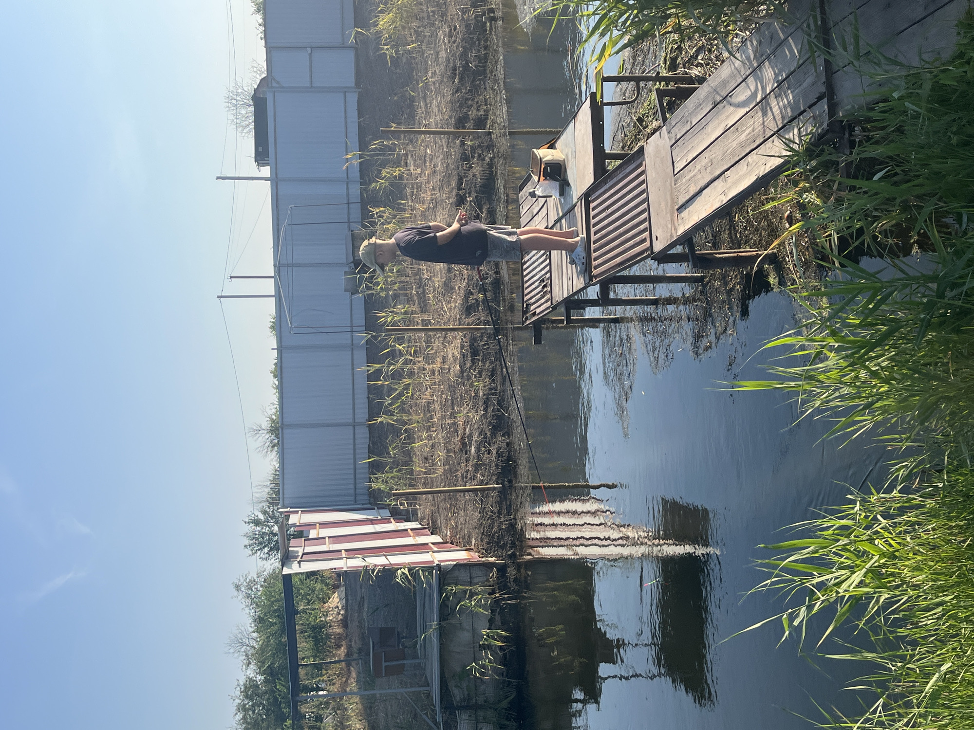 A young male, wearing a cap, dark t-shirt, and patterned shorts, stands on a wooden pier fishing in a pond. He is facing away from the camera, looking towards the water. The pier is made of weathered wood and metal grating, supported by rusty metal poles. On the pier, next to the male, is a small tackle box and a water bottle. The pond water is dark and reflects the sky and surrounding structures. The background features a white metal fence, some vegetation, and a building with red and white siding. Tall green grass grows along the edge of the pond in the foreground. The sky is clear and blue. The scene appears to be in a rural or semi-rural setting.