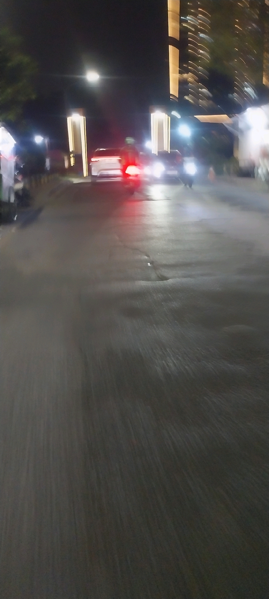 A nighttime street scene in Bencongan Indah, Indonesia, captured with motion blur. In the middle ground, a white car with illuminated taillights is visible, along with a motorcycle, also with blurred lights. Two tall, illuminated vertical structures are positioned on either side of the road. In the background, a tall building with many illuminated windows is blurred. The asphalt road is wet and reflects the light from the vehicles and structures. Trees are visible on the left side of the image, and a streetlight is prominent in the upper left quadrant. The overall impression is a dynamic urban environment at night, with traffic and illuminated architecture.
