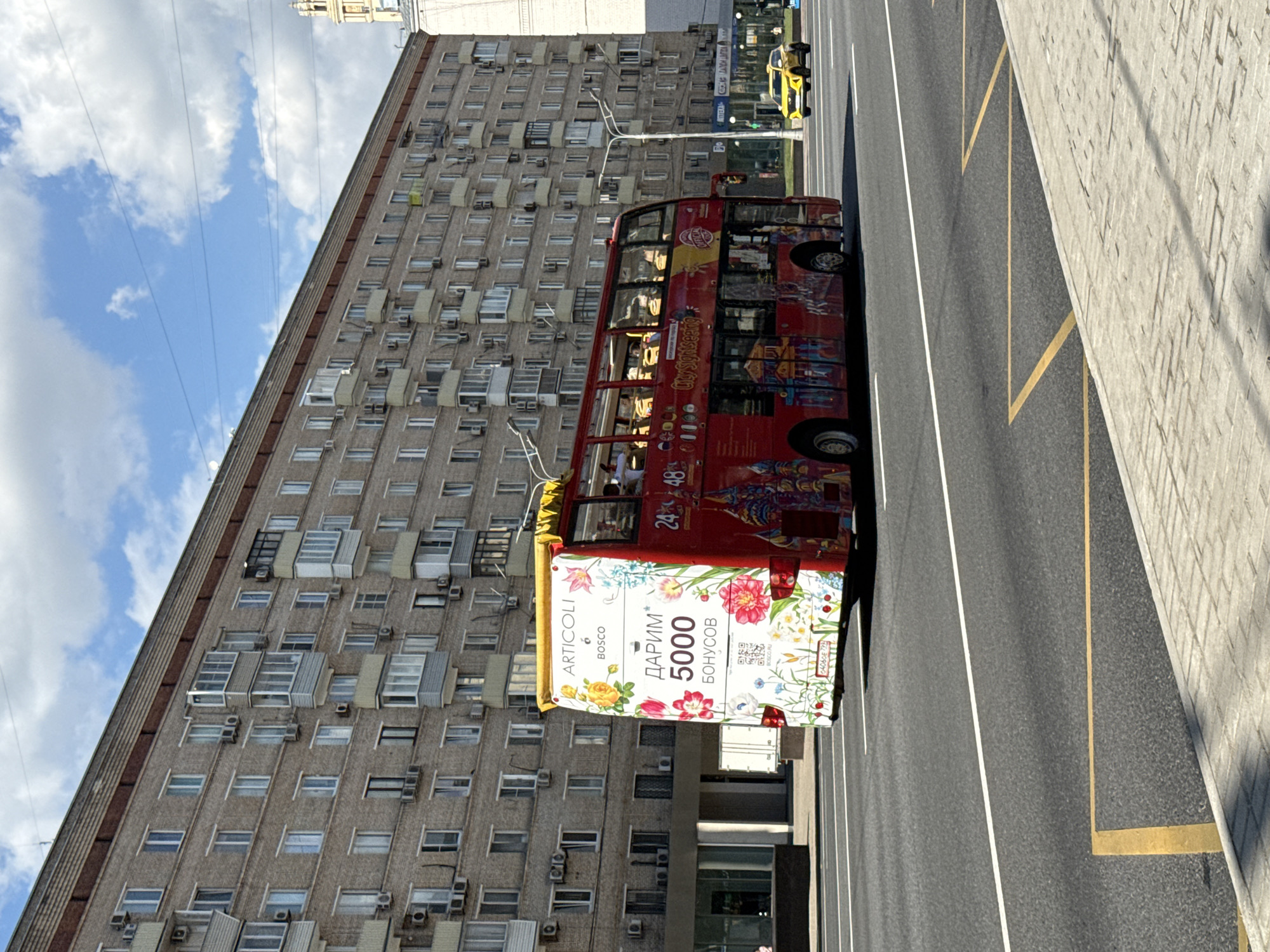 The image shows a red double-decker tour bus driving on a street in Moscow, Russia. The bus has a floral design on the back with Russian text that says "DARIM 5000 BONUSOV," which translates to "We give 5000 bonuses." The bus is decorated with a colorful image of what appears to be a Kremlin-like structure. In the background, a tall apartment building with many windows and air conditioners is visible. A yellow car is also seen driving on the street. The sky is blue with white clouds. The street is paved with asphalt, and there are white and yellow lines marking the lanes. The scene is set during daytime.