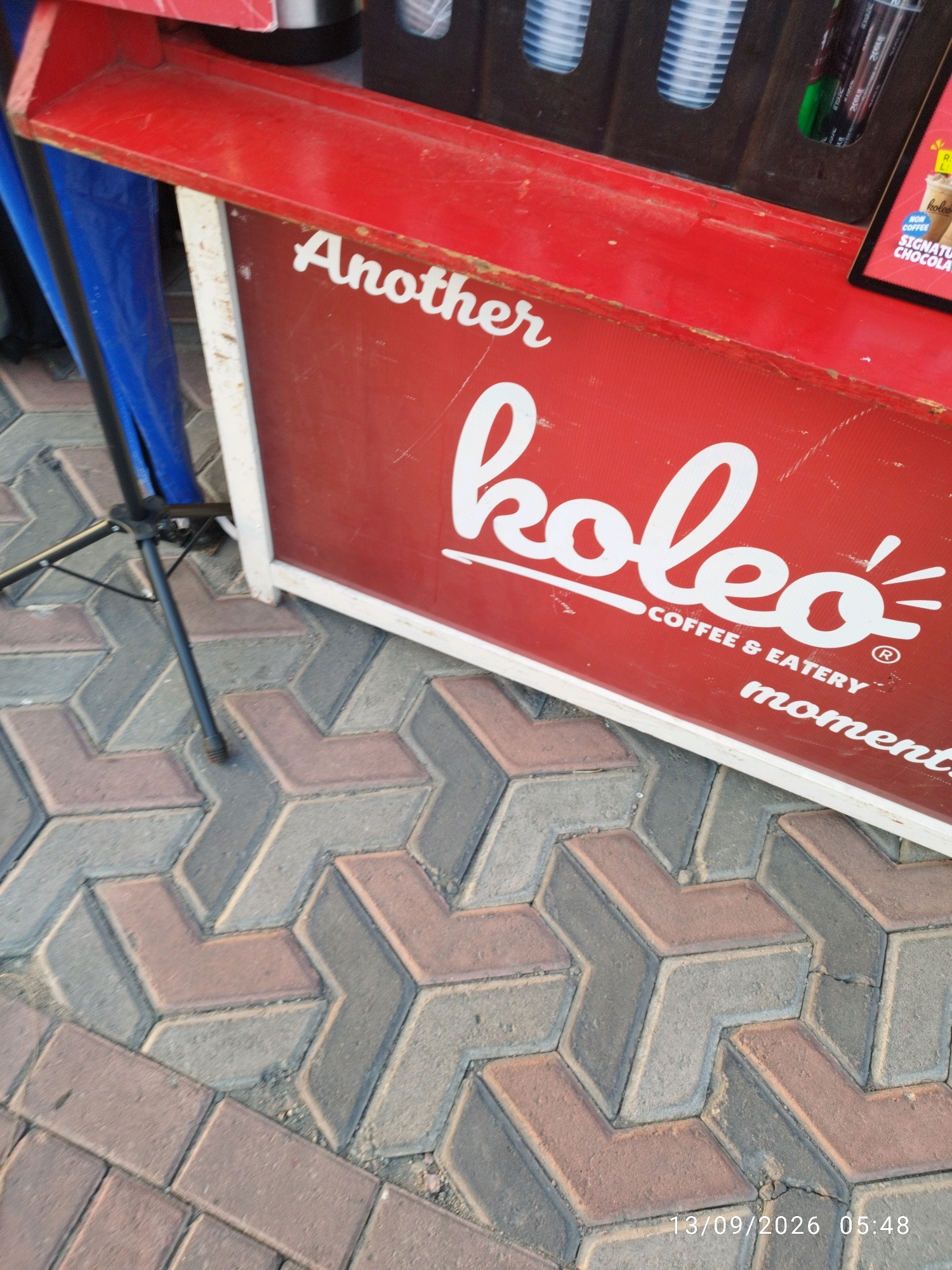 The image shows a red sign with white text that says "Another koleo COFFEE & EATERY moments". The sign is mounted on a red structure and is framed by a white border. To the left of the sign, a blue tarp and a black tripod are visible. The ground is paved with interlocking bricks in a chevron pattern, in shades of grey and reddish-brown. A date and time stamp "13/09/2026 05:48" is visible in the lower right corner of the image. The scene appears to be outdoors, likely on a sidewalk or public space. The location is identified as Mojokerto, Indonesia. There are no people visible in the image.