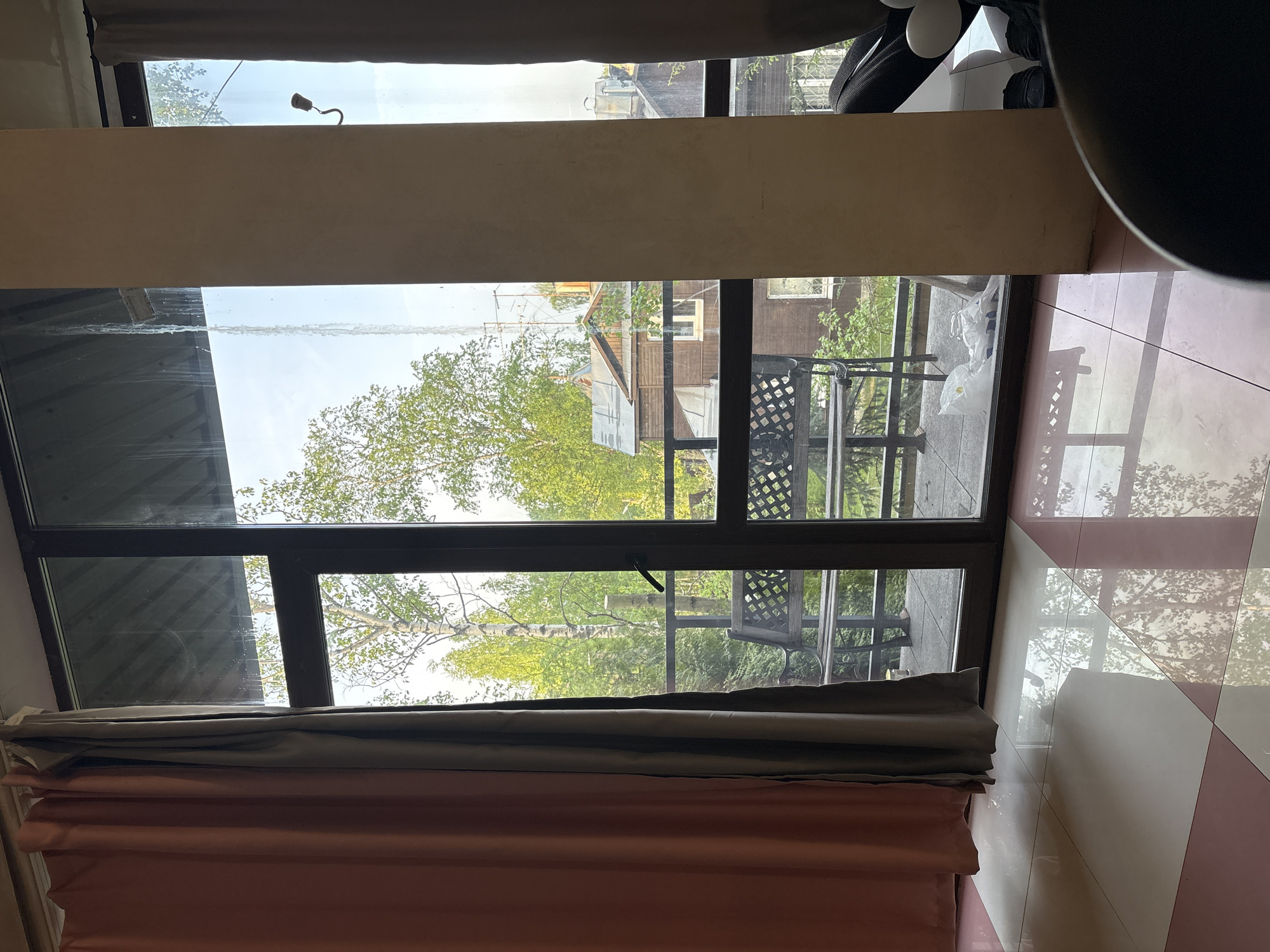 The image captures an interior view through a window, offering a glimpse of the outside world. The window frame is dark, contrasting with the light seen through the glass. Outside, there are trees with fresh green foliage, suggesting springtime. A structure resembling a building or house with a balcony is visible. The balcony has lattice chairs and furniture. A tiled surface of the floor is visible with pink and white tiles.
Given the visible architectural style and natural environment, it is plausible that the location is in a residential area of Saint Petersburg, Russia, known for its architecture and green spaces.