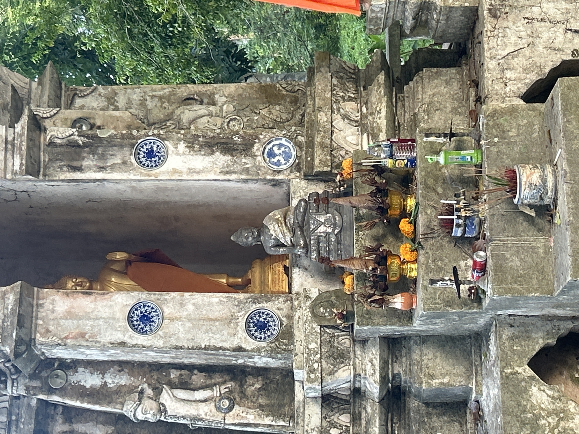 The image depicts a Buddhist shrine located in Ko Pha-ngan District, Thailand. In the center of the shrine, a golden Buddha statue stands behind a smaller, seated stone Buddha statue. The shrine is adorned with decorative blue and white ceramic plates, as well as offerings of flowers and incense. The structure itself is made of aged stone, showing signs of weathering and wear. Surrounding the shrine are lush green trees, indicating an outdoor setting. Several smaller statues or carvings are visible on the stone facade. At the base of the shrine, various items are placed, including incense sticks in a pot, a red and white can, and several bottles, suggesting recent offerings or ongoing rituals.