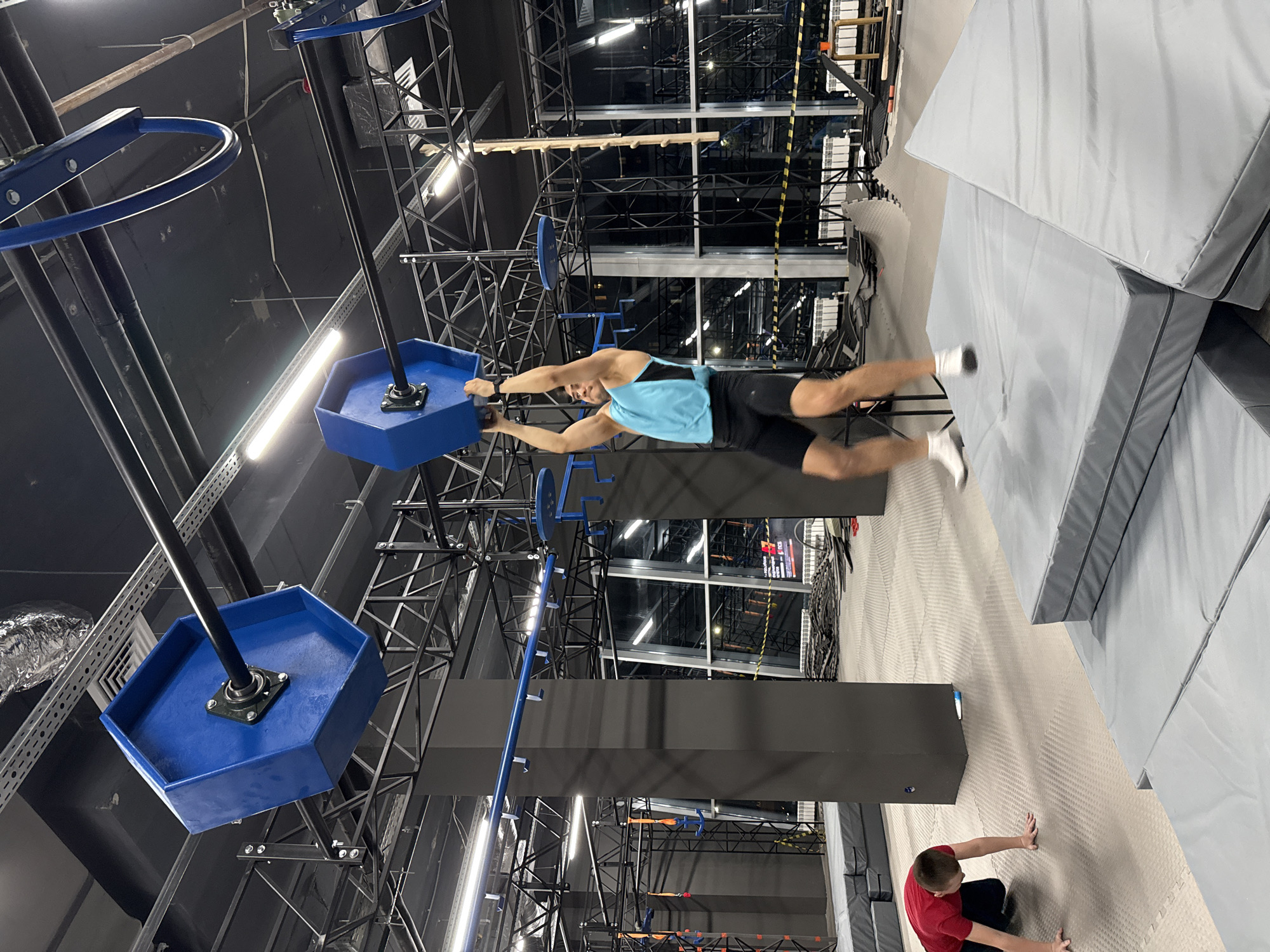 An athletic young man is captured mid-air in an indoor obstacle course, likely a ninja warrior or parkour gym. He is suspended horizontally, gripping a blue hexagonal platform with both hands, his body outstretched and legs blurred with motion, suggesting a powerful swing or leap. He wears a light blue and black sleeveless top with black shorts and white socks.
The gym features a complex overhead structure of black metal trusses and beams. From this structure, several blue hexagonal platforms on black poles hang down, forming part of an agility challenge. Another similar platform is visible further to the left. The floor is covered with light gray, textured padding, while large, light and dark gray safety mats are stacked to the right, indicating a landing zone. In the bottom right corner, another young man in a red t-shirt and black pants is on his hands and knees on the padded floor, looking up towards the person in the air, perhaps observing or awaiting his turn.
The setting is an industrial-style indoor facility with exposed pipes, ventilation ducts, and bright artificial lighting from fluorescent tubes. Through windows in the background, more of the facility's interior or an external view with indistinct lights is visible, along with a blurry screen displaying illegible text or graphics. The overall ambiance is dynamic and focused on physical challenge, with the time of day being indeterminate due to the indoor lighting. The image's perspective is rotated 90 degrees clockwise.