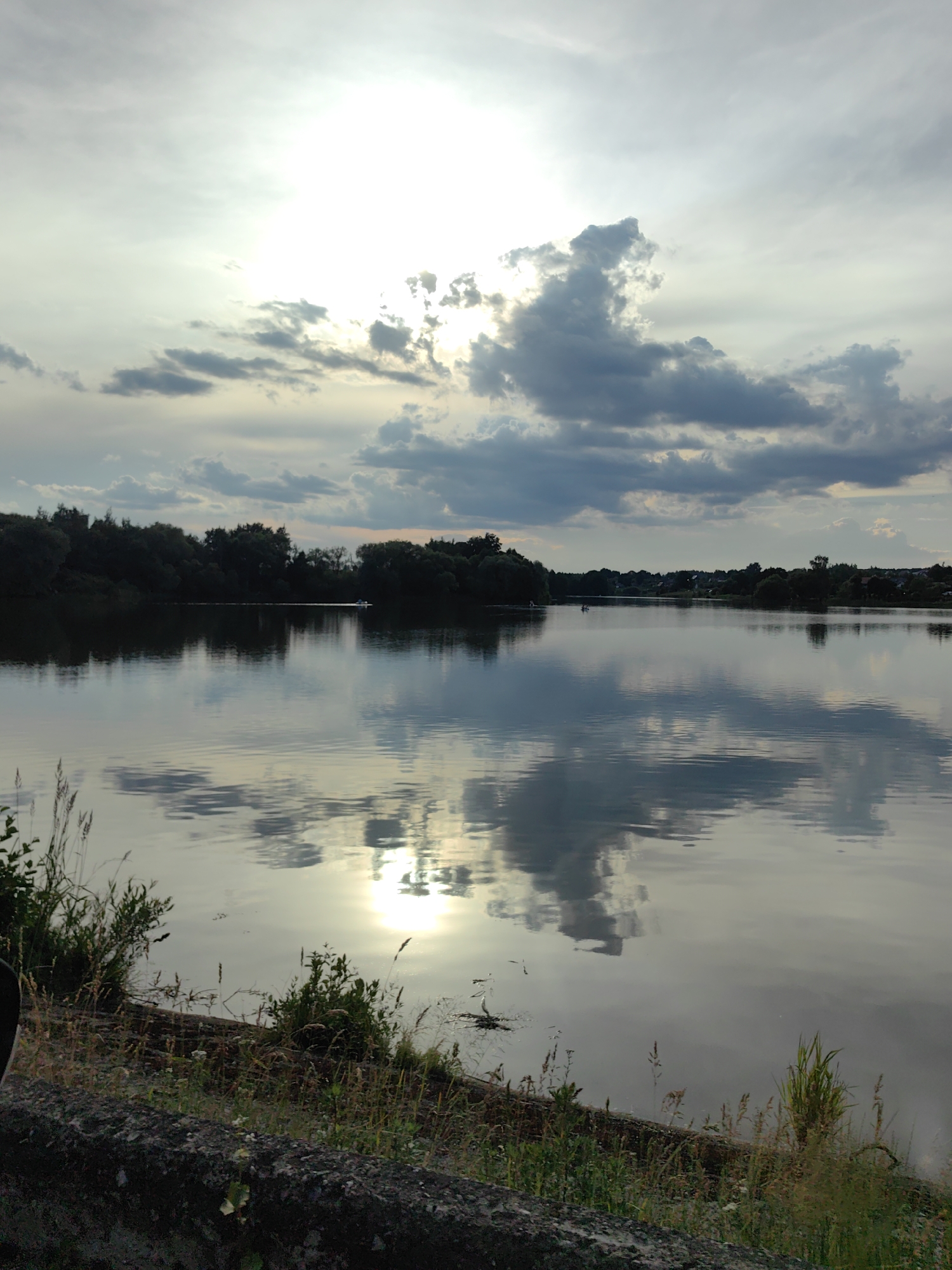 The image shows a wide, calm body of water, likely a lake or river, reflecting the sky and surrounding trees. The sky is partly cloudy with the sun partially obscured, creating a bright, diffused light. Dark, cumulus clouds are visible, particularly in the upper right portion of the sky, and their reflections are clearly seen on the water's surface. Along the far bank of the water, a dense line of dark green trees creates a silhouetted horizon. In the distance, small structures, possibly houses or buildings, are visible interspersed with more trees. A very small, light-colored boat with at least two occupants is moving across the water in the mid-distance, leaving a faint wake. The foreground features a low, textured concrete barrier with scattered dry grasses and some green vegetation growing along its edge. The overall impression is a tranquil, natural landscape.
