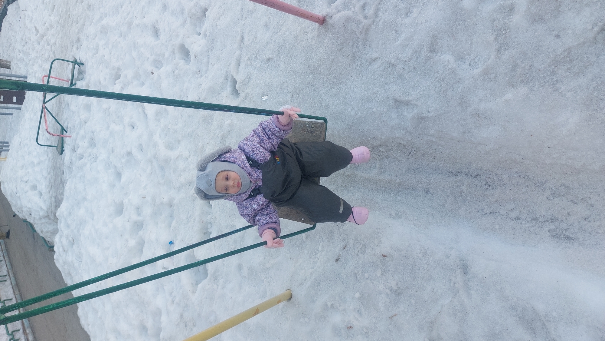 A child wearing a winter hat, a purple and pink jacket, and dark pants with pink boots is on a swing in a snowy outdoor setting. The swing is made of green metal with a wooden seat. Surrounding the swing are patches of snow. A glimpse of other playground equipment is visible in the background, including some red and green structures. Given the presence of snow and the clothing of the child, the photo was taken during the winter months. The exact location within the city of Novosibirsk, Russia cannot be determined from this image.