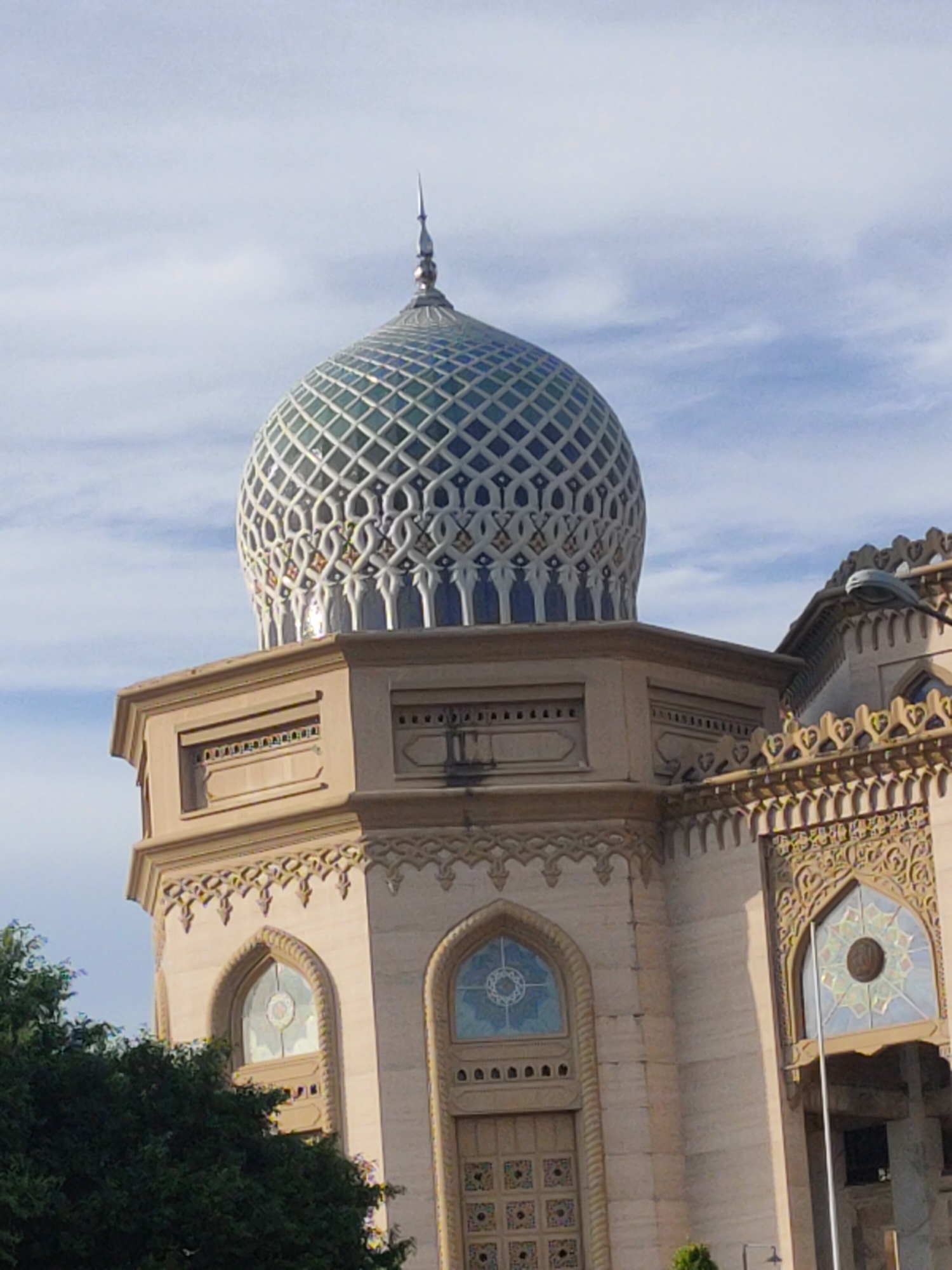The image displays a close-up view of an ornate mosque, likely located in Lhokseumawe, Indonesia, characterized by its distinctive architectural style. The central focus is a large, elaborate dome that crowns the structure. The dome features a captivating geometric pattern, appearing to be constructed from tiles or mosaic in shades of blue, light blue, and white or light grey, and is topped with a slender, pointed finial.
The building's facade beneath the dome is constructed from a light-colored, possibly sand-colored or beige, material. This section includes decorative moldings and smaller, rectangular openings directly below the dome. Further down, multiple arched windows are visible, adorned with intricate stained glass displaying geometric or star-like patterns. The building's exterior is richly decorated with elaborate carved patterns and borders, showcasing traditional Islamic architectural motifs along cornices and around window frames.
The sky in the background is a clear, light blue with scattered, wispy white clouds, indicating daytime. Green foliage from trees is visible in the lower-left corner and partially in the lower-right, framing the base of the structure. No people or specific activities are depicted in the image, emphasizing the mosque's architectural grandeur. There is no visible text.