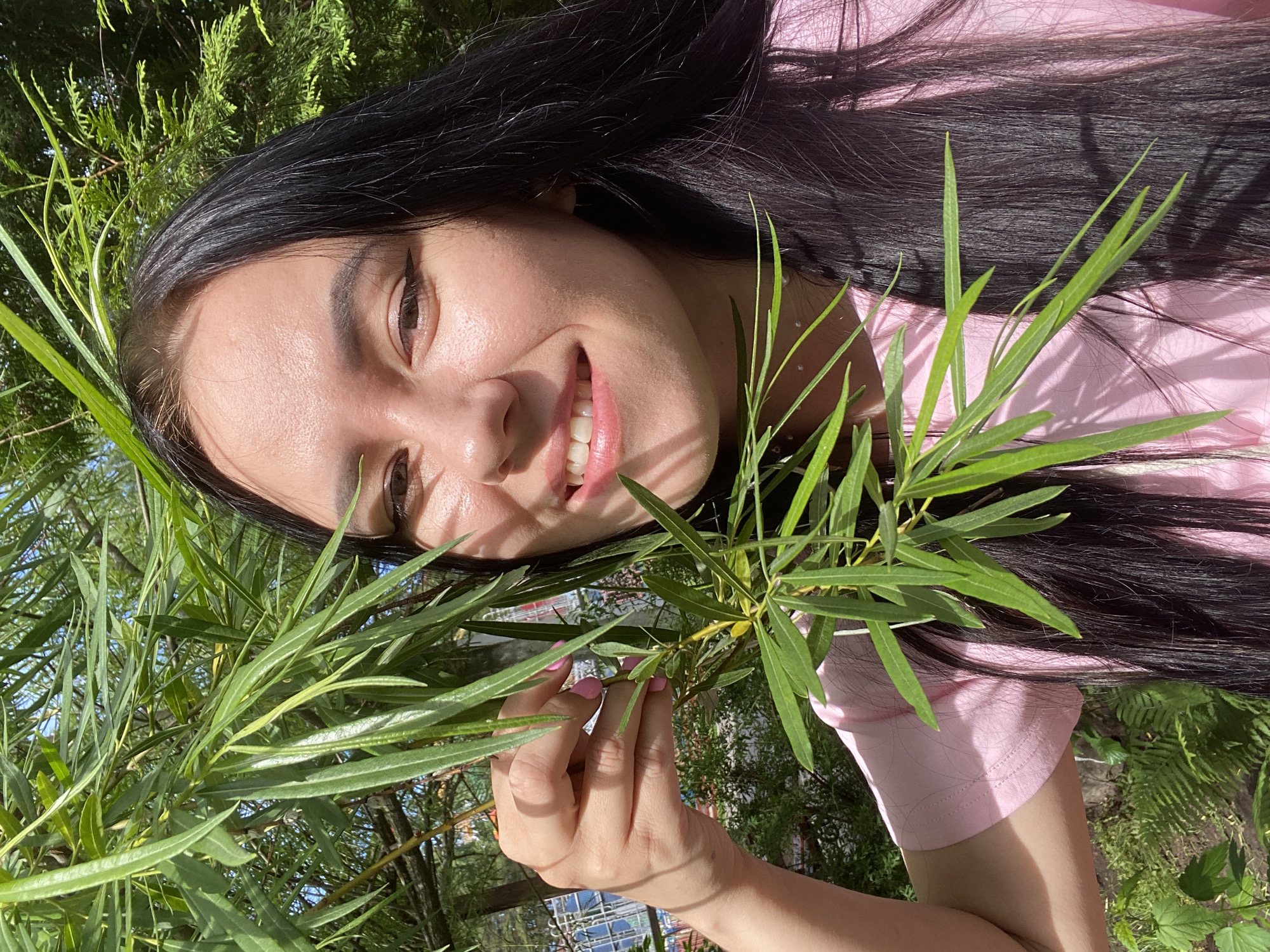 A young woman, possibly of East Asian descent, is pictured outdoors amidst lush green foliage under bright, direct sunlight. She is positioned at an angle, with her head towards the left side of the frame, smiling broadly towards the camera. Her long, dark hair cascades around her head and upper body. She is wearing a light pink, short-sleeved top.
One of her hands, with nails painted pink, gently holds a branch with slender, vibrant green leaves that extend across the center of the image, partially obscuring her and the background. Her dark eyes are visible, enhanced with winged eyeliner. Her expression conveys happiness and contentment.
The background consists of blurred green trees and bushes, suggesting a park, garden, or natural environment. Strong shadows are cast by the leaves onto her hair and clothing, indicating a clear, sunny day, likely morning or afternoon. A faint glimpse of a structure or fence can be seen in the distant background through the foliage. The overall atmosphere is cheerful and tranquil. There is no visible text in the image.