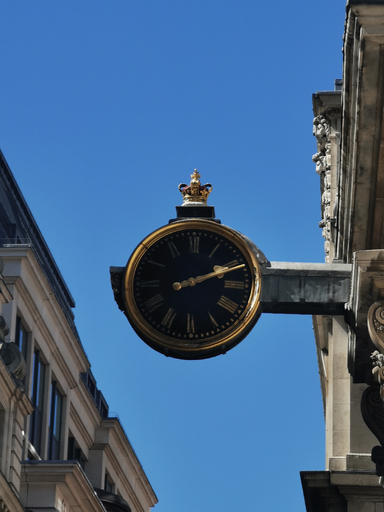 A grand, circular street clock with a black face and golden Roman numerals (I through XII) is the central focus of the image, set against a vibrant, clear blue sky. The clock's golden hands indicate the time is approximately 10:45. Crowning the timepiece is an ornate, golden miniature crown, adorned with what appear to be red and blue jewels, suggesting a royal or official connection.
The clock is suspended by a sturdy, dark grey metal bracket extending from a richly detailed building on the right. This building features elaborate architectural carvings, including what seem to be gargoyle-like figures and decorative stone work, characteristic of historical urban architecture. On the left, a different building frames the scene, distinguished by lighter-colored stone or concrete, multiple windows with dark frames, and a visible dark roofline.
The absence of clouds and the bright illumination suggest a sunny morning. The combination of the classic architectural styles and the prominent crown on the clock strongly places the scene in London, United Kingdom, known for its regal heritage and iconic street furniture. No people or specific events are visible, allowing the intricate details of the clock and surrounding buildings to stand out against the expanse of the sky.
