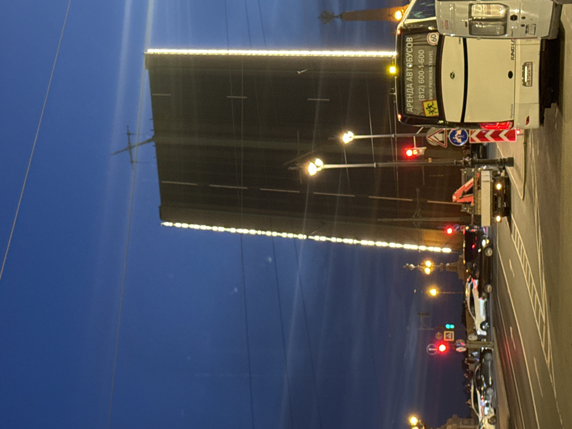 This photograph captures a nighttime or late twilight scene in Saint Petersburg, Russia, focusing on a raised bascule bridge. The sky is a deep blue with faint, horizontal cloud streaks, indicative of the "White Nights" phenomenon common in Saint Petersburg during summer. The central subject is a large, dark bascule bridge with its two main leaves raised high into an almost vertical position, creating a significant gap for maritime traffic. The inner edges of these raised bridge leaves are brightly illuminated with vertical strips of warm white or yellow LED lights, reflecting on the slightly textured surface of the bridge structure. In the upper right background, a tall, slender column with a distinct finial or sculpture at its apex is partially visible, characteristic of Saint Petersburg's Rostral Columns. Overhead, thin electrical or communication wires crisscross the sky. In the foreground and midground, a multi-lane road is visible, marked with white solid and dashed lines, as well as a pedestrian crossing. Numerous vehicles are present: * On the right, the rear of a white King Long tour bus is prominently featured, displaying the text "АРЕНДА АВТОБУСОВ" (Bus Rental), a Saint Petersburg phone number "(812) 600-1-600," and "WWW.PREMIERA.TRAVEL." A circular "60" speed limit sign is affixed to its rear. To its immediate right, the front portion of a white van is visible. * To the left, several cars are stopped or moving on the road, including a white SUV, a black sedan, and other darker vehicles. * Further left, a light-colored flatbed truck with a small crane attachment is parked or moving slowly. The scene is illuminated by numerous artificial light sources: * Multiple streetlights with yellow-orange glowing globes line the road and bridge structures. * Traffic signals are visible; one on the left displays a red light, while another shows a green signal in the distance. * Various road signs are present, including red circular "no entry" or "prohibited" signs, a blue circular "mandatory turn right" sign, and triangular warning signs with red borders. * The bridge's support structures and decorative elements are partially visible on the left, illuminated by scattered lights. No specific individuals are clearly discernible. The overall impression is an urban environment during an evening of significant activity, centered around a functional and aesthetically illuminated bascule bridge, strongly identified as the Palace Bridge due to the (812) area code on the bus and the presence of the Rostral