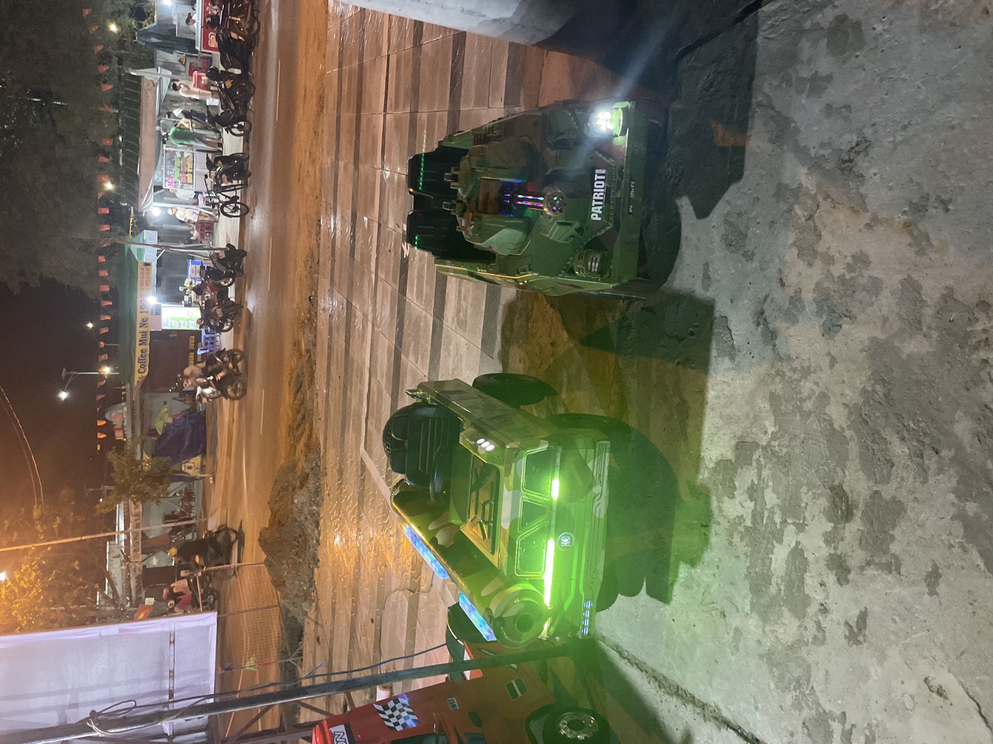 The image depicts a nighttime scene in Mũi Né, Vietnam. The location seems to be an urban area with a street lined with businesses and parked motorbikes. The background shows the street is well-lit with streetlights. There's a row of street vendors or shops visible on one side of the road, and the area appears bustling with activity.
In the foreground, there are two children's toy vehicles positioned on a concrete surface, likely a sidewalk or pedestrian area. One is a miniature tank-like vehicle with the word "PATRIOT" displayed on its side. The other one is a camouflage-patterned truck. The toys are lit with bright, colorful LED lights. These toy vehicles appear to be part of a recreational setup, possibly for children to ride.