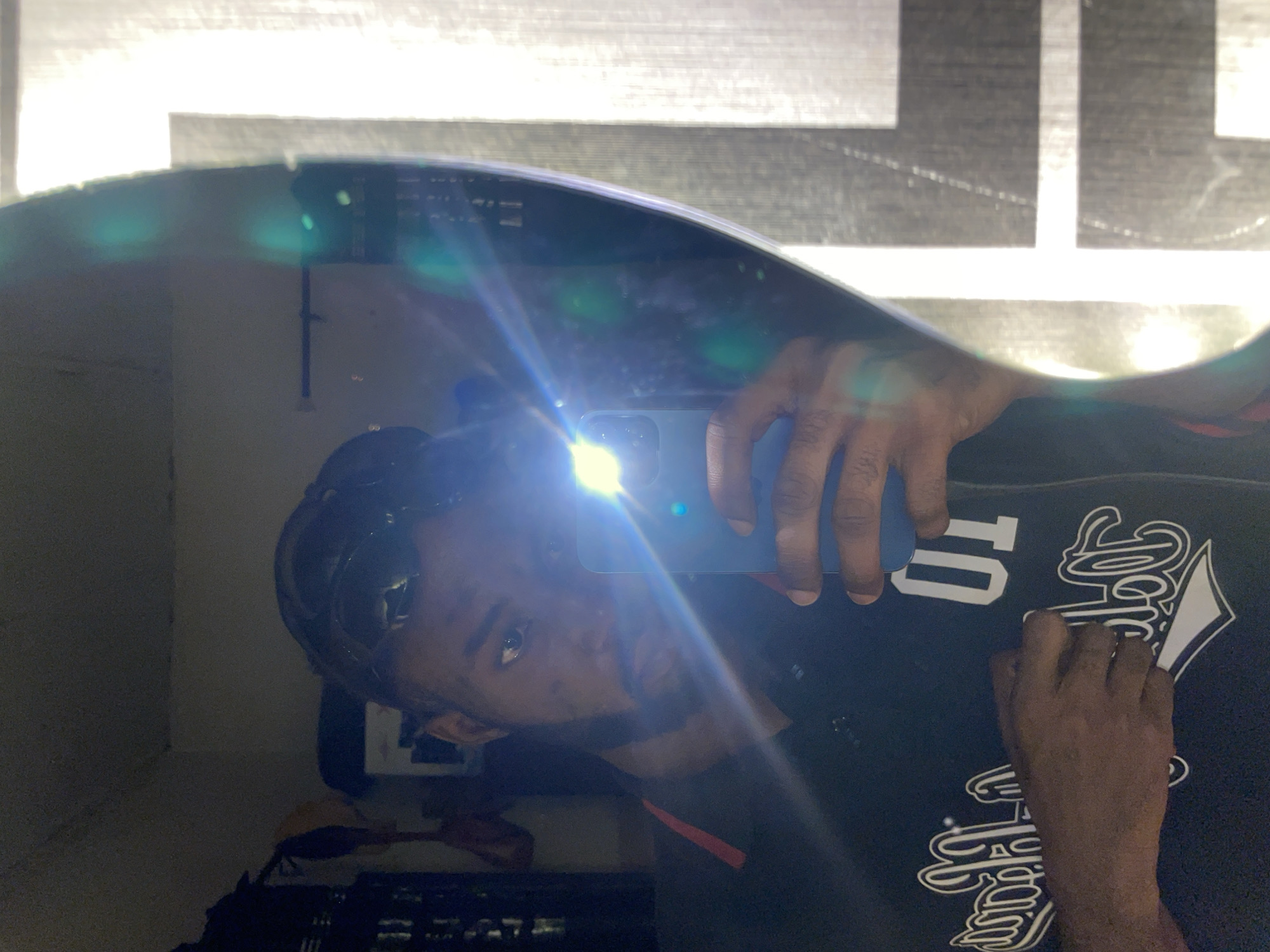 A male individual is captured in a mirror selfie taken indoors in Lokoja, Nigeria. He presents as a person of African descent, with short dark hair and a beard. He is wearing a dark polo shirt with a red accent strip along the placket and a white graphic design on the chest that includes stylized text and the number "01". His right hand holds a dark-colored smartphone, which is angled towards the mirror, its activated flash producing a strong bright light and significant lens flare that traverses the frame. His left hand is positioned over his shirt. The mirror reflects a dimly lit room, with a dark furnishing unit visible on the left side of the reflection. The right side of the mirror's reflection shows a brightly illuminated vertical section, suggestive of a window with blinds or a textured wall, contrasting with the interior's lower light levels. The mirror itself exhibits a slightly irregular or curved edge on the right.