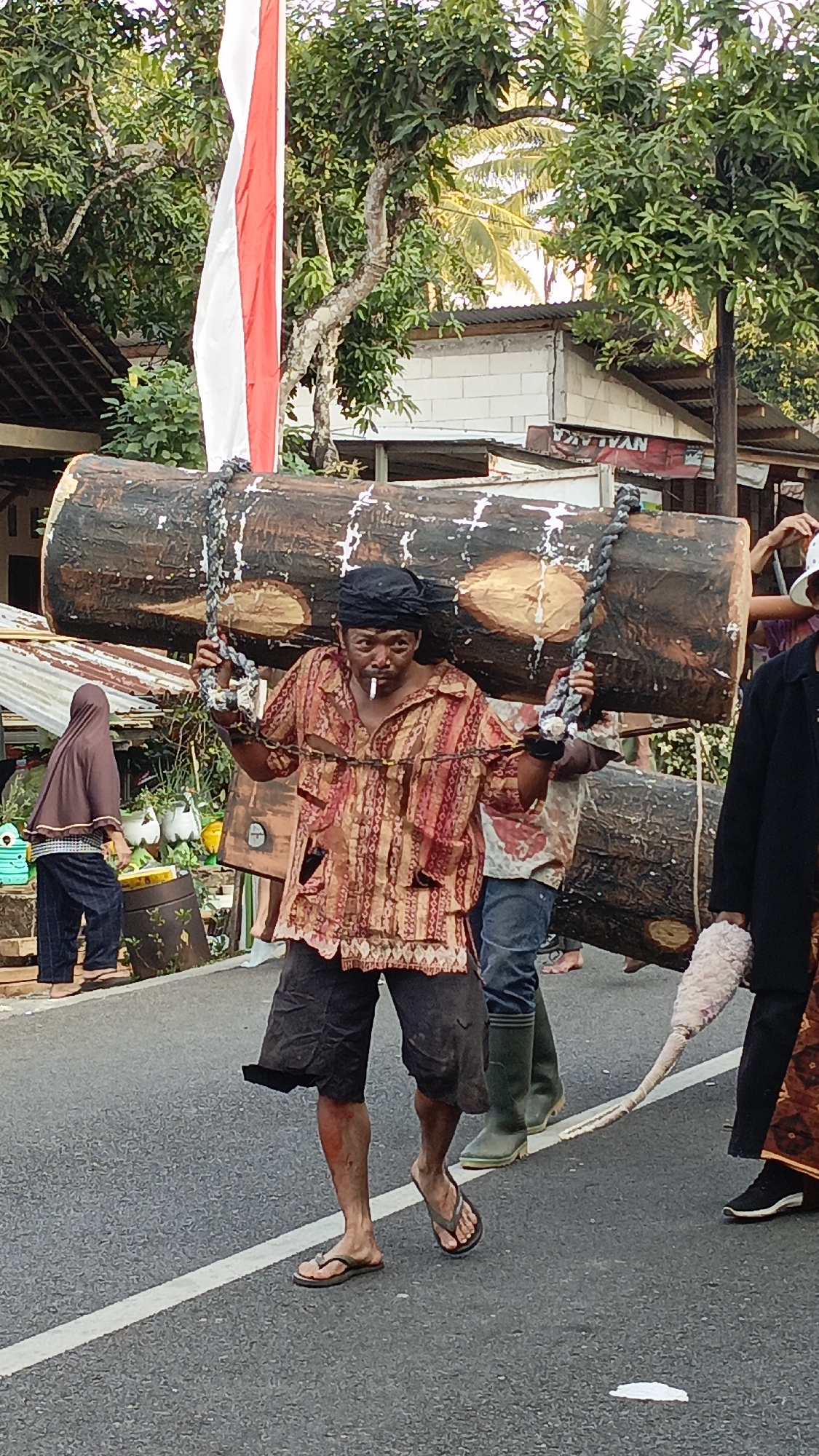 In the city of Krajan Lor, Indonesia, a group of men participate in a procession, carrying a large, thick log supported by ropes and chains. The lead man, wearing a black headscarf, a vibrantly patterned shirt, and dark shorts, balances the log on his shoulders while smoking a cigarette. His companions, with their faces obscured, are also carrying the log from behind. The event takes place on a paved road, with a white line marking the lane. In the background, a red and white Indonesian flag flutters above lush green trees and modest buildings, suggesting a festive or ceremonial occasion. The scene is illuminated by natural sunlight, casting soft shadows and highlighting the textures of the log and the clothing of the participants.