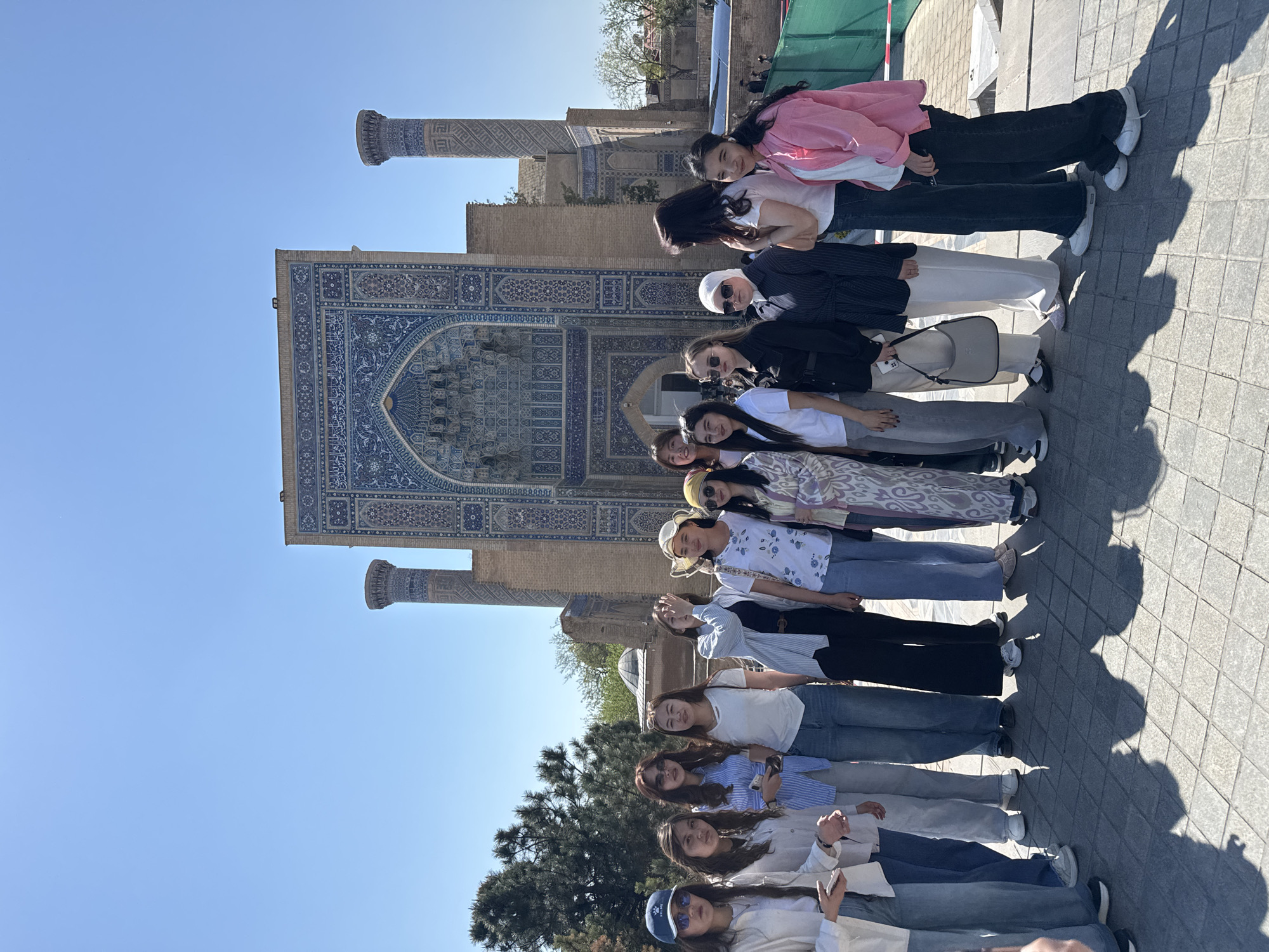 A horizontal, eye-level, outdoor shot shows a group of 13 young women standing in a line perpendicular to a large blue and white tile building. The women are all standing close together, most with their arms down. The women are dressed in a variety of styles. At the top of the group (furthest away) the woman is wearing a pink shirt. The woman at the bottom of the group has on a blue hat. The large building behind them is made of white and blue tiles. On the right and left, just in front of the building are two matching white pillars decorated with the same tile as the building. In the background, in the center of the image is some sparse greenery. The ground around the group is made of gray square stones. The sky is clear and blue. The location is Samarkand City, Uzbekistan.