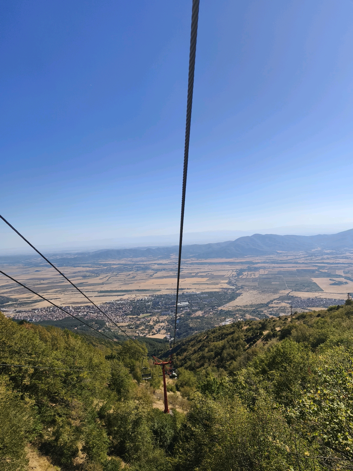 An aerial, eye-level, full shot captures a ski lift descending a lush, green mountainside. The ski lift cables stretch across the clear, blue sky, with one cable prominently featured in the foreground, running vertically from the top to the bottom of the frame. Below, a red ski lift tower supports a single ski lift seat, occupied by two individuals. The mountainside is covered in dense green trees and foliage. In the background, a sprawling city with red-tiled roofs is nestled in a valley, surrounded by arid, brown fields and distant, hazy mountains. The scene is bathed in bright daylight, suggesting a clear, sunny day.