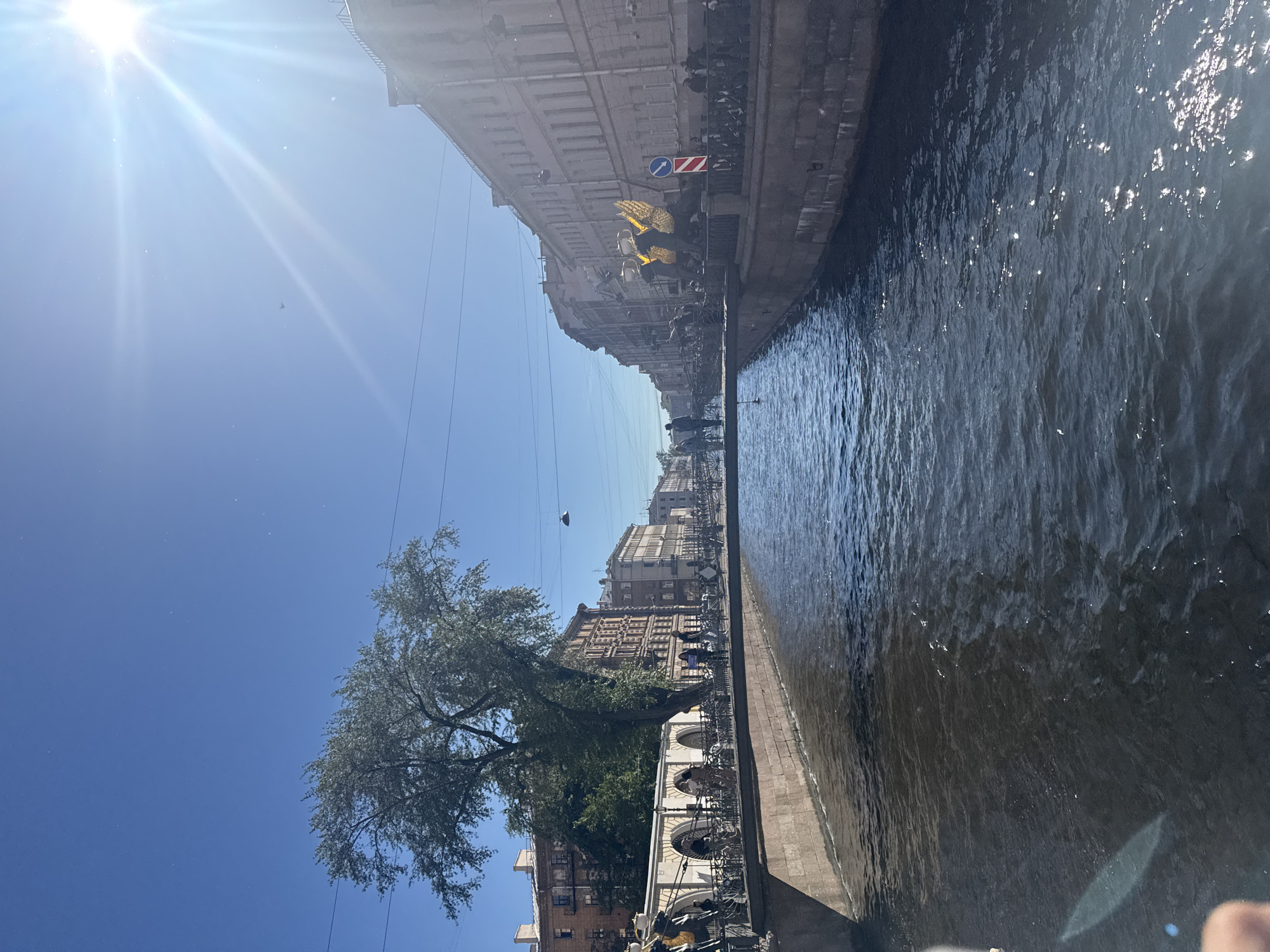 The image captures a sunny day along a canal in Saint Petersburg, Russia. The canal, with dark, rippling water, is lined by stone embankments. A pedestrian bridge spans the canal, with people walking and standing on it. Bicycles are parked along the bridge's railing. On the left bank, a large tree with full foliage stands near an arched walkway. Buildings with light-colored facades and numerous windows line the right bank, extending into the distance. The sky is a clear, bright blue, with the sun prominently positioned on the right side of the frame, casting lens flare. A small airplane is visible in the sky. The overall scene suggests a lively urban environment with both historical architecture and recreational activity.