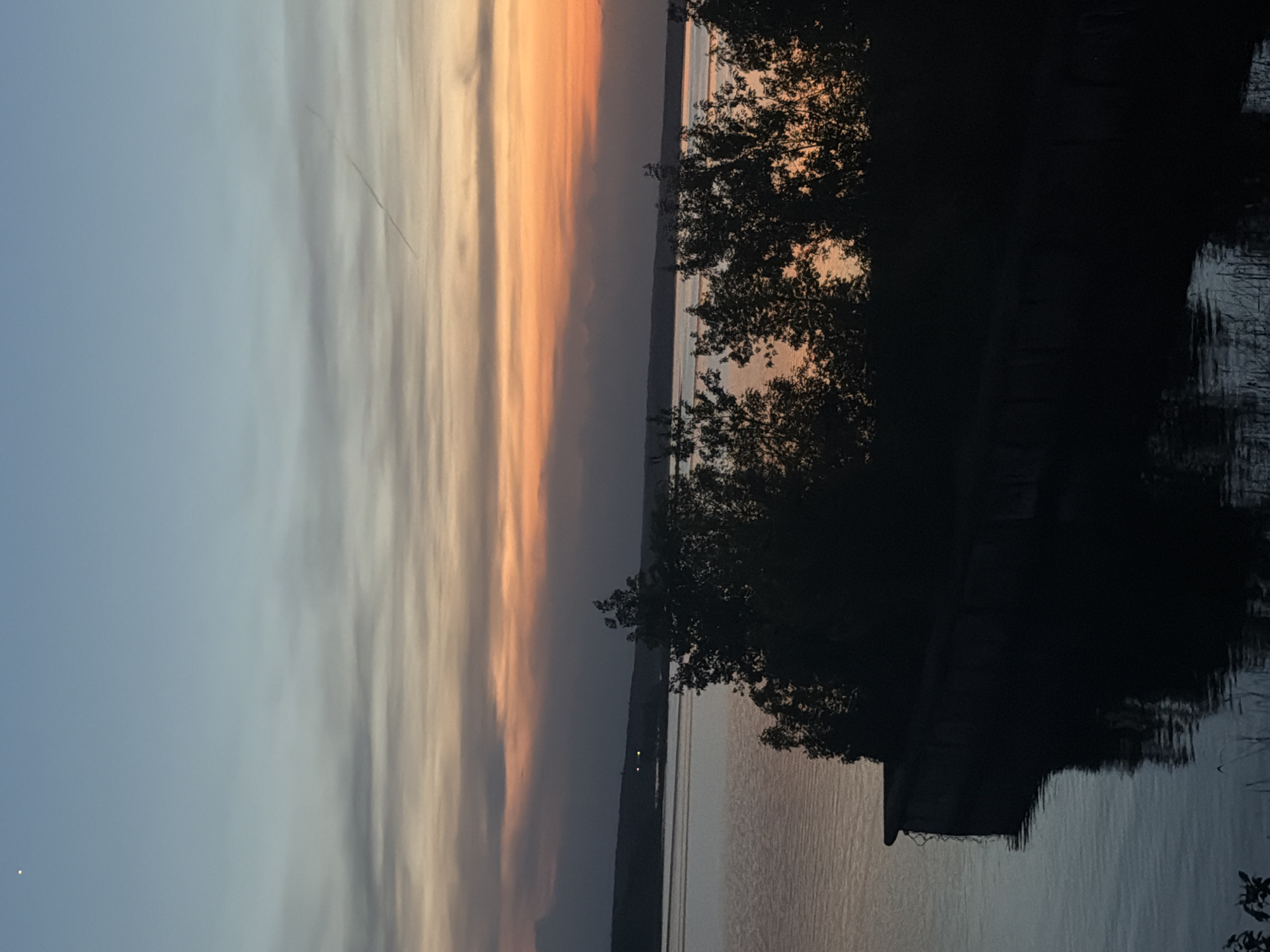 The image captures a sunset over a body of water in Izhevsk, Russia. The sky transitions from a pale blue on the left to a warm orange-red gradient on the right, with striated cloud formations. A contrail from a plane stretches across the upper portion of the sky. A line of trees and a low structure are silhouetted against the water and the vibrant sunset. The water is calm with subtle reflections of the sky's colors and the dark silhouettes of the trees and structure.