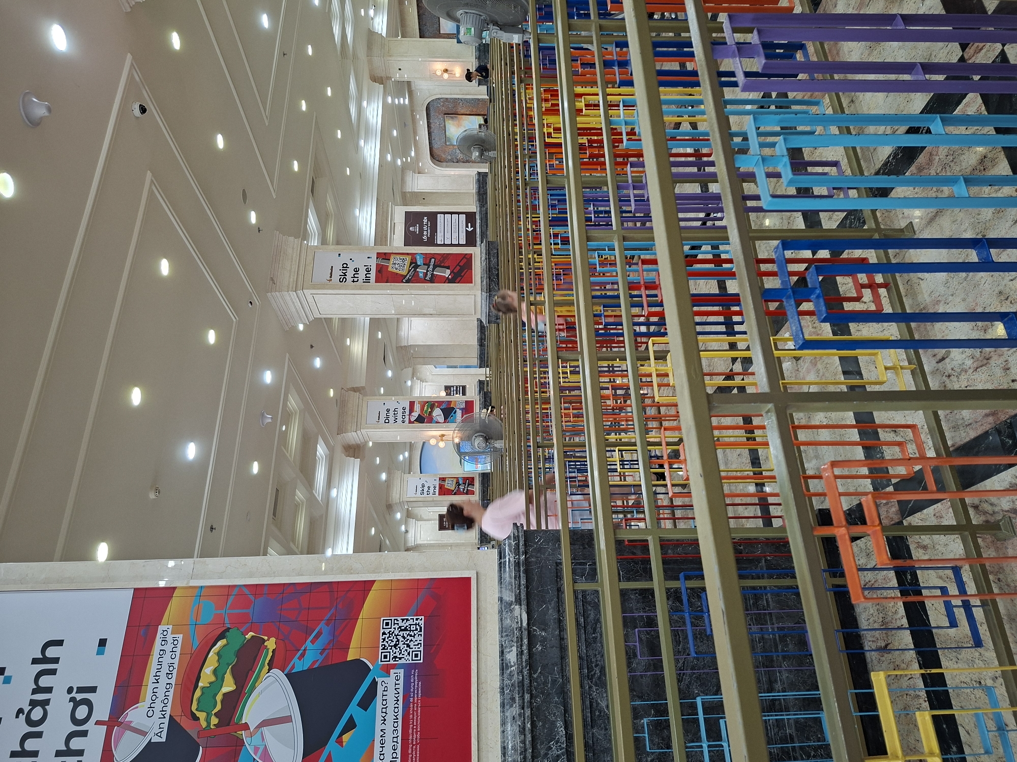 The image depicts the interior of a commercial space, possibly a shopping mall or a large store in Nha Trang Ward, Vietnam. The scene is viewed from a slightly upward angle, emphasizing the height and depth of the space.
The foreground is dominated by a colorful, abstract geometric structure, potentially a decorative railing or art installation. It features square and rectangular shapes in a spectrum of colors, including yellow, orange, red, blue, and purple.
Behind the colorful structure, the interior reveals a grand hallway with a high, intricately designed ceiling featuring recessed lighting. The walls are lined with pillars and promotional displays. One poster is prominently visible, advertising food with accompanying text in Vietnamese and Russian.
Several ceiling fans are visible, suspended from the high ceiling. People are sparsely scattered throughout the space, one wearing a pink shirt and another further down the hall. The scene suggests a bustling, commercial environment.