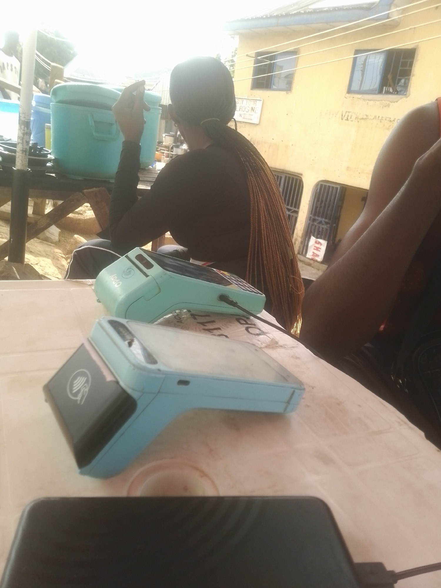 The image shows two light blue OPay point-of-sale devices on a white table. One device is closer to the viewer and shows a contactless payment symbol. The other device is further away and has a screen. In the background, a person with braided hair is seated at the table, with their back to the camera. They are wearing a black long-sleeved shirt. To the right of this person, another person's arm and shoulder are visible, wearing an orange top. Behind the table, there is a turquoise container, and further back, a yellow building with windows and a tiled roof. The scene appears to be outdoors, possibly in a market or informal business setting. The lighting suggests it is daytime. There is some writing visible on the building in the background, but it is mostly illegible. One sign appears to say "BUY YOUR..."