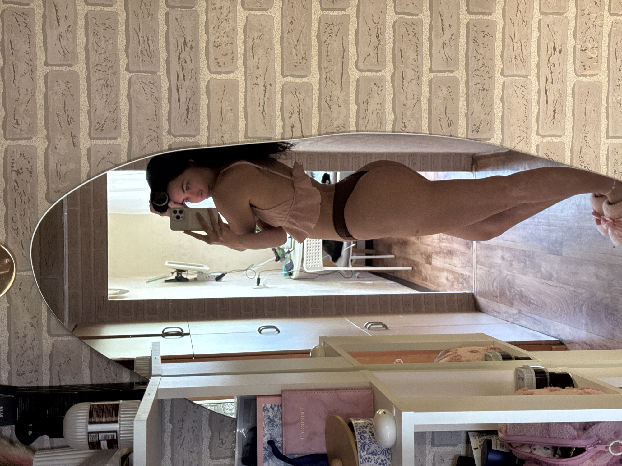 The image shows a young woman standing in what appears to be a bedroom or dressing area. She is wearing a light pink ruffled top and brown underwear. She is holding a smartphone in her right hand, taking a mirror selfie. Her hair is styled with rollers. The room has brick wallpaper and a wooden floor. To the left of the woman is a white shelving unit with various items, including books, bags, and containers. A large, oval mirror reflects the woman and part of the room. There is a doorway visible in the background, leading to another area with a table and chair. The overall impression is that of a casual, personal space.