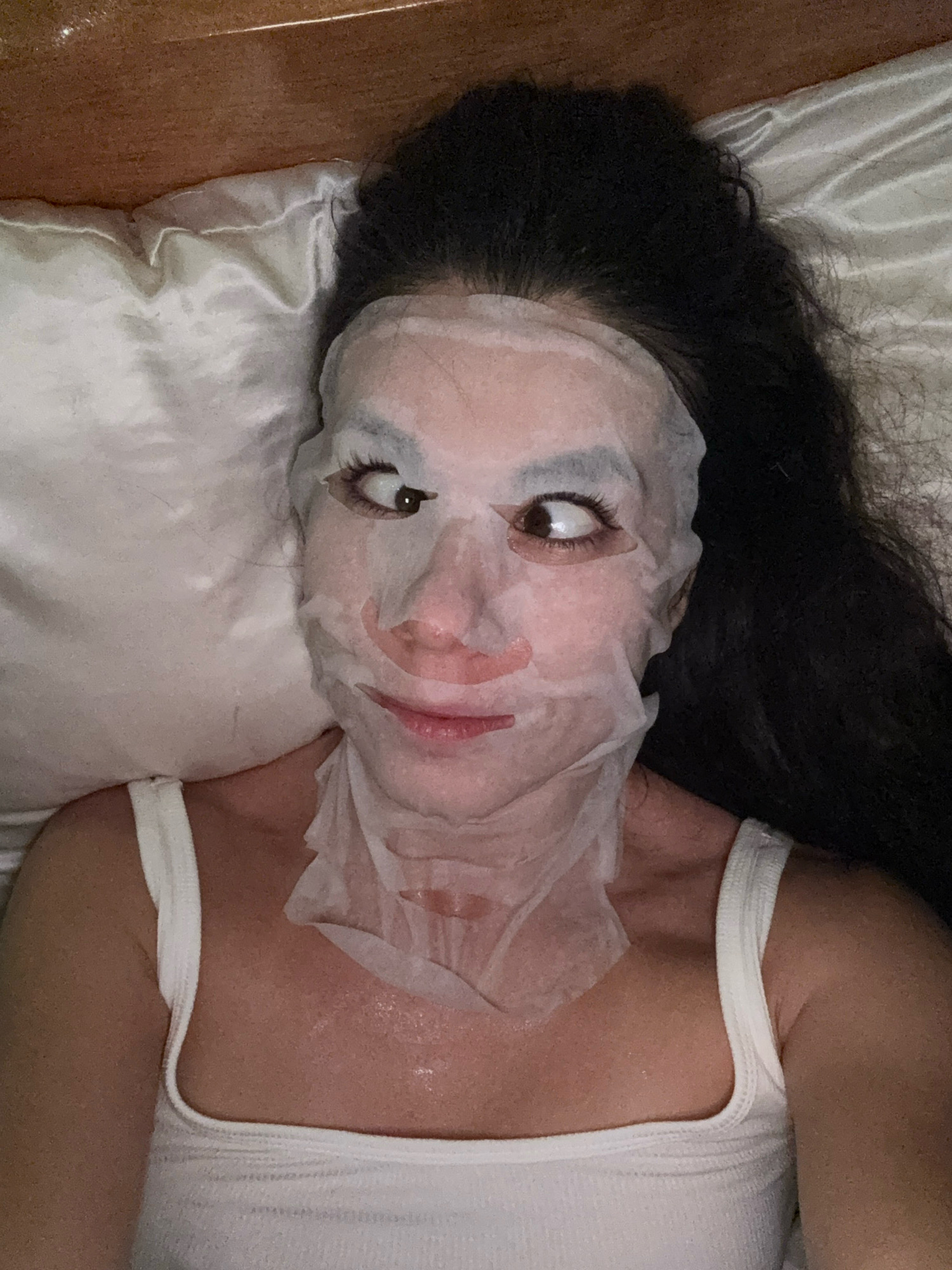 A woman with dark hair lies on a satin pillow and bedding, wearing a white tank top and a sheet mask that covers her face. Her eyes are crossed, and she has a slight smile. The scene is lit by artificial light, and the background is a wooden headboard. The image is taken from a high angle, looking down at the woman.