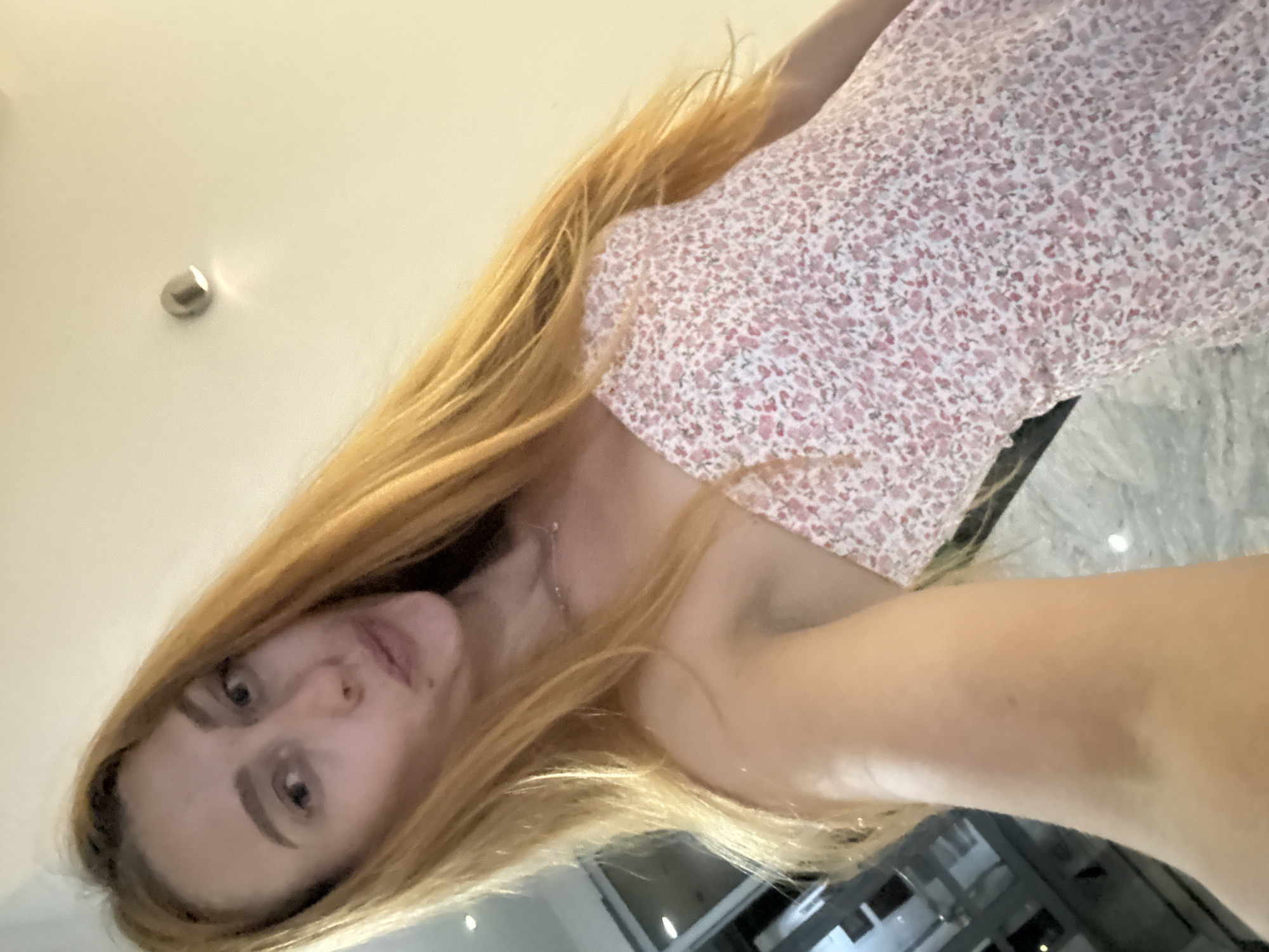 A woman with long, blonde hair and fair skin is seen in a selfie. She is wearing a strapless, floral-print dress with a pink and white pattern. The background is dimly lit and appears to be indoors, with a light fixture visible on the ceiling. Based on the provided context, the location is identified as Budva, Montenegro. The woman's face is turned slightly, and she is looking towards the camera with a neutral expression. Her left arm is visible, holding the camera. The lighting suggests an indoor environment, possibly a hotel room or apartment.