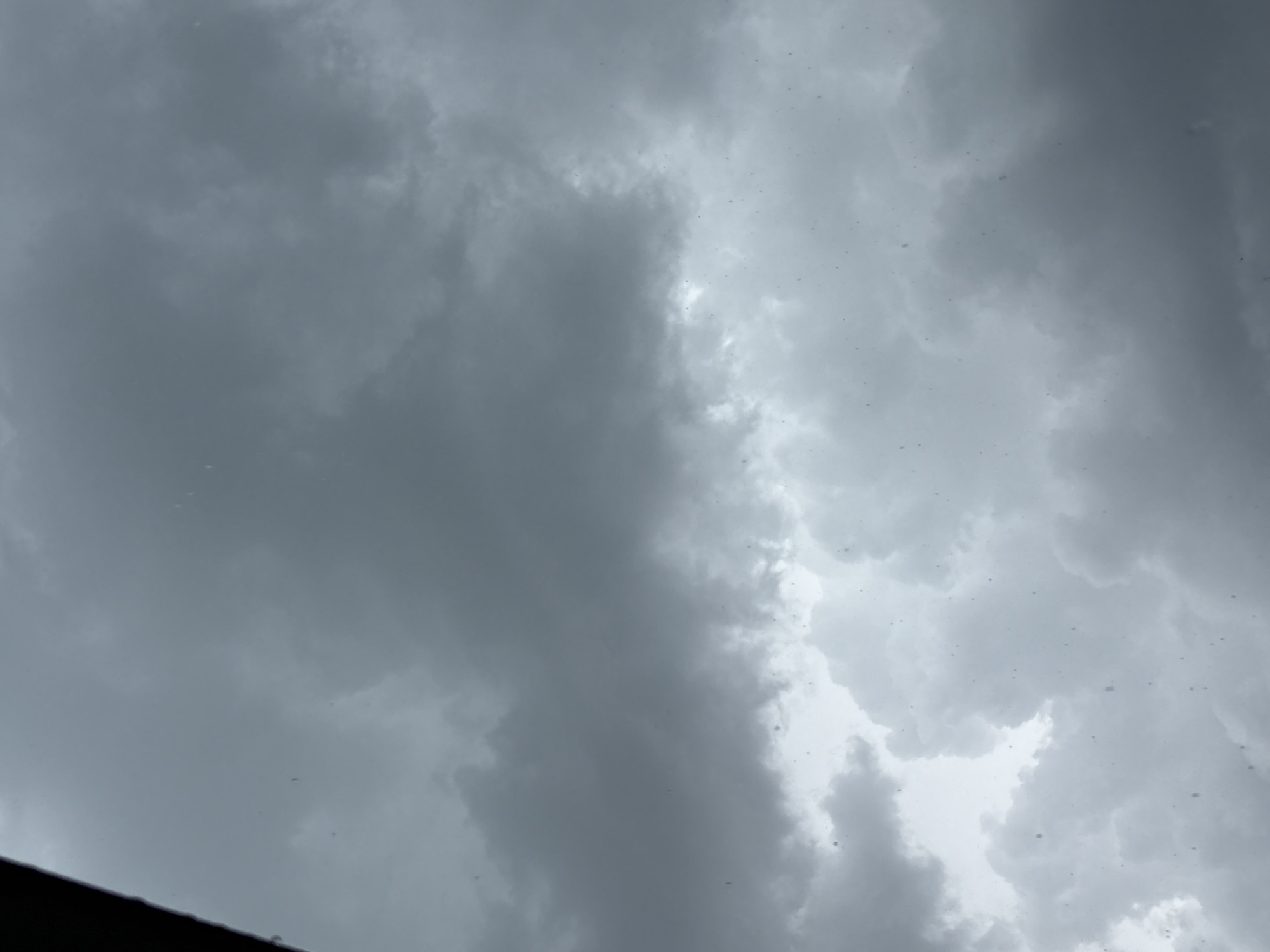 The image displays a heavily overcast sky above Plovdiv, Bulgaria, dominated by dense, multi-layered grey clouds that vary from dark leaden tones to lighter grey-white. Numerous small, irregularly shaped specks, consistent with water droplets on a transparent surface, are scattered across the frame, indicating rain or condensation. A dark, indeterminate structural element partially encroaches from the upper-left corner. The scene is illuminated by dim, diffused light, characteristic of inclement or heavily overcast weather conditions. No human figures or specific ground-level objects are discernible within the sky depiction.